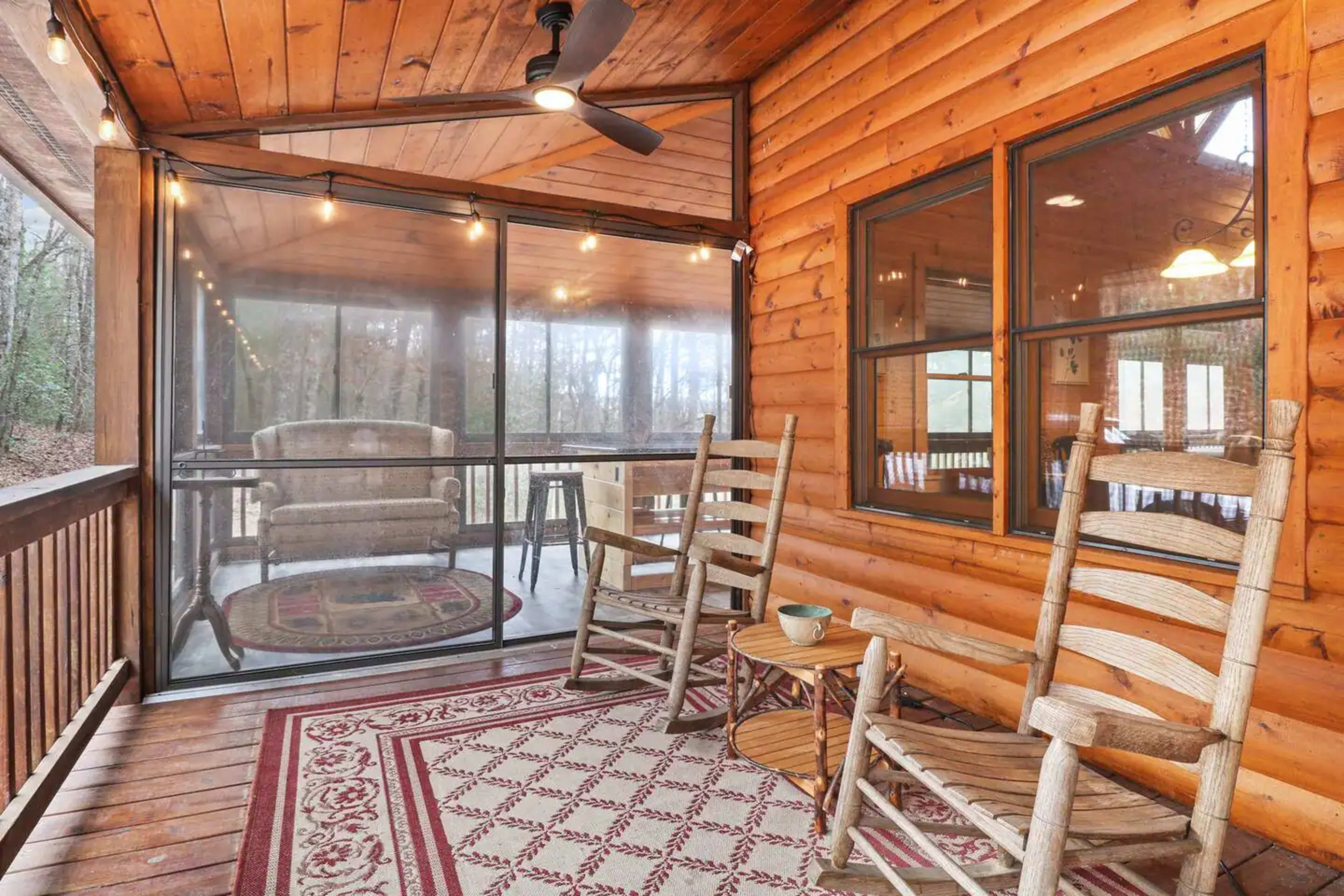 A screened porch with wooden rocking chairs, a rug, and large windows overlooking a forested area.