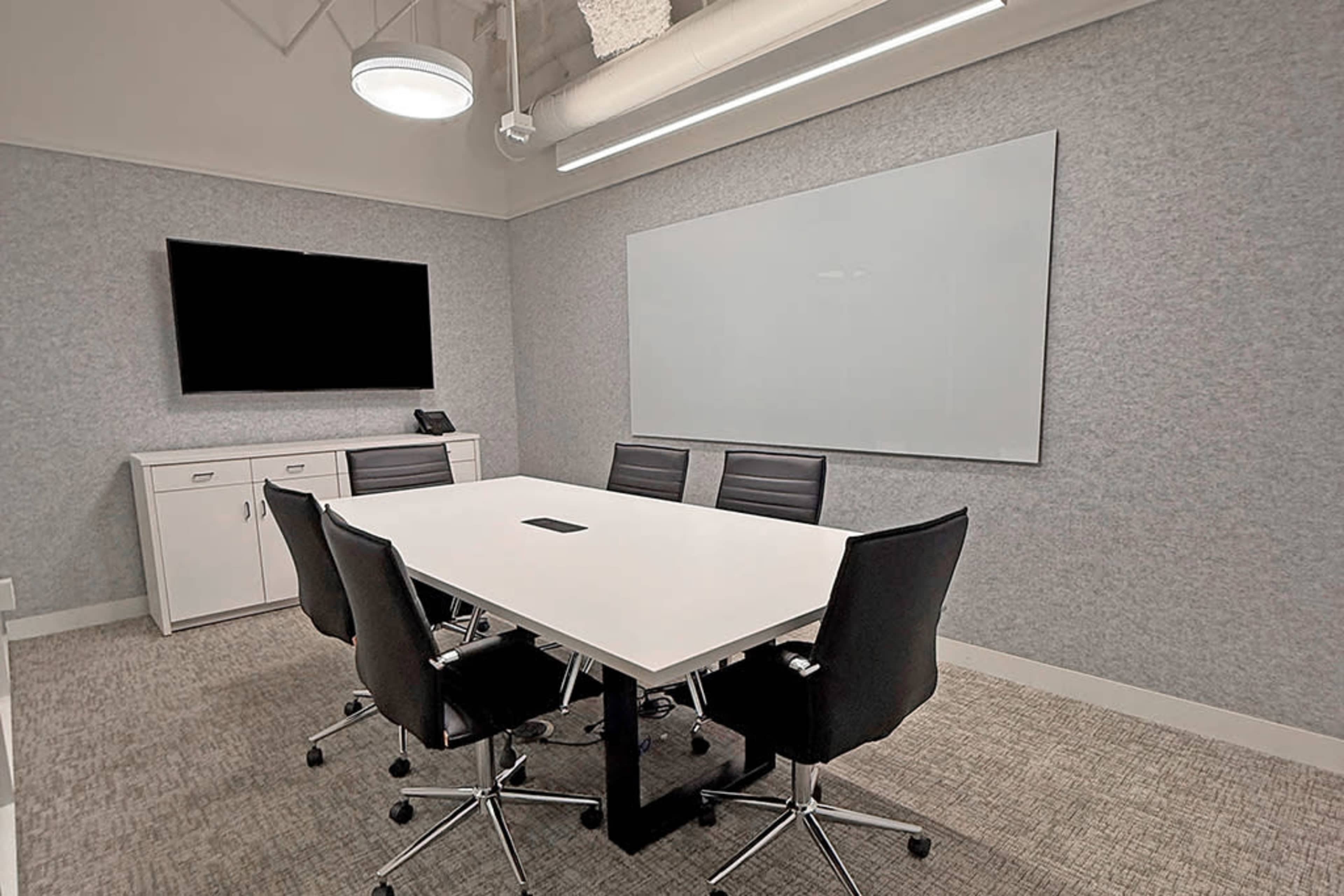 The image shows a modern conference room featuring a white rectangular table surrounded by black rolling chairs, with a large screen on one wall and a whiteboard on another.