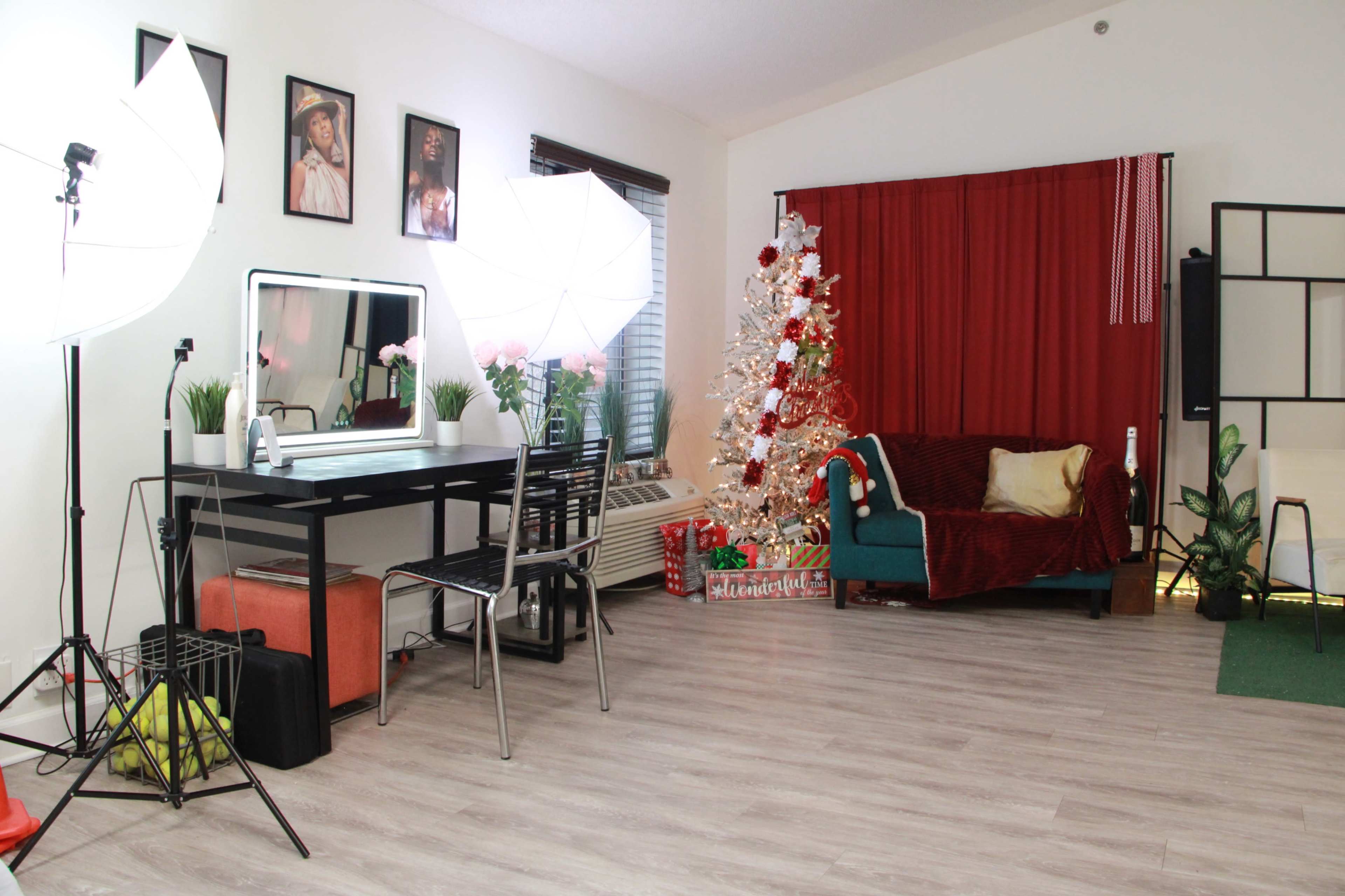 The image shows a bright room featuring a decorated Christmas tree, a teal sofa with red throw pillows, a table with a mirror and studio lighting equipment, and a red curtain background.