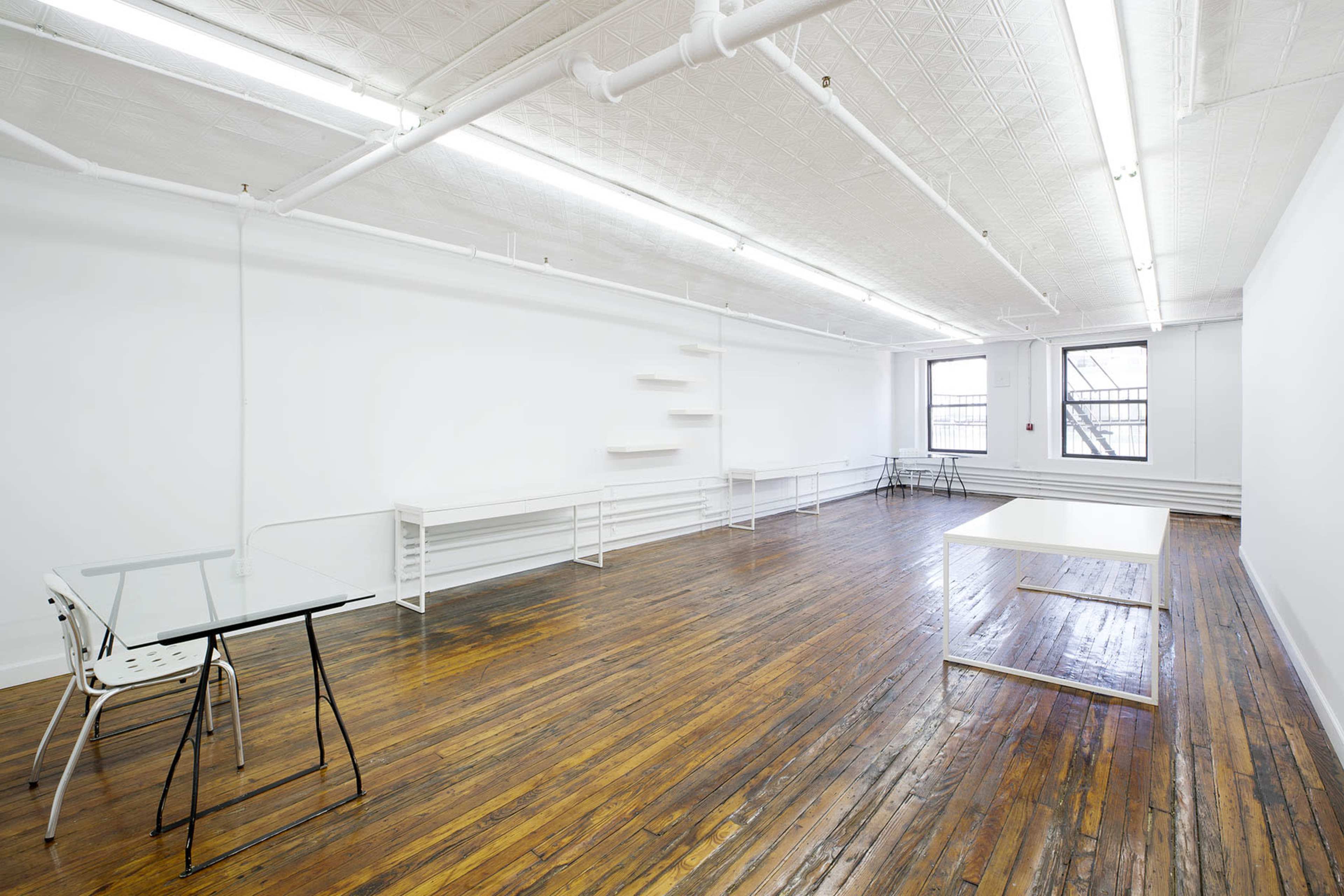The image shows a spacious, empty room with wooden floors, white walls, and minimal furniture including a glass-top table and white shelves.