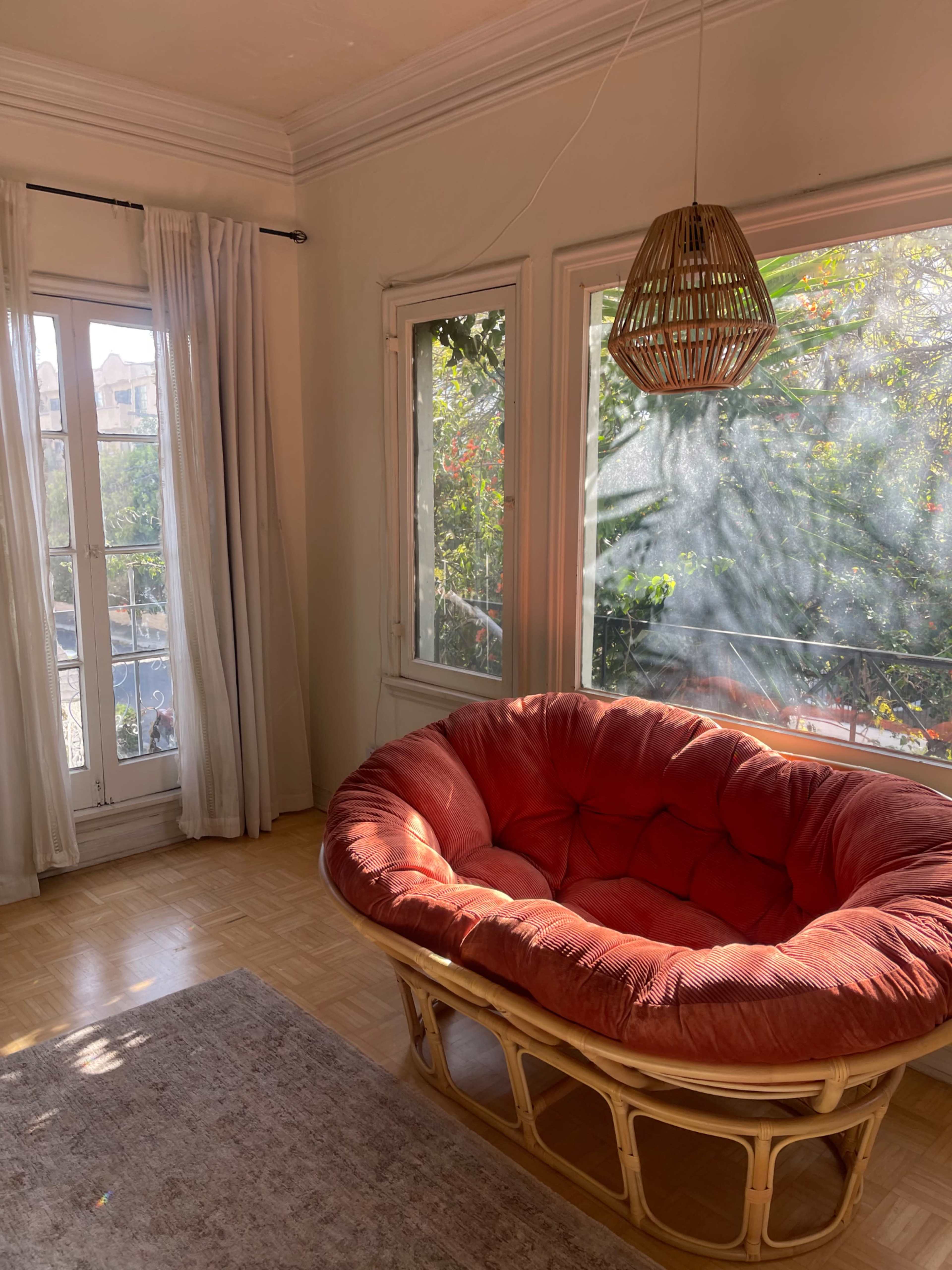 A large, round, red cushioned chair sits in a brightly lit room, with windows dressed in white curtains and greenery visible outside.