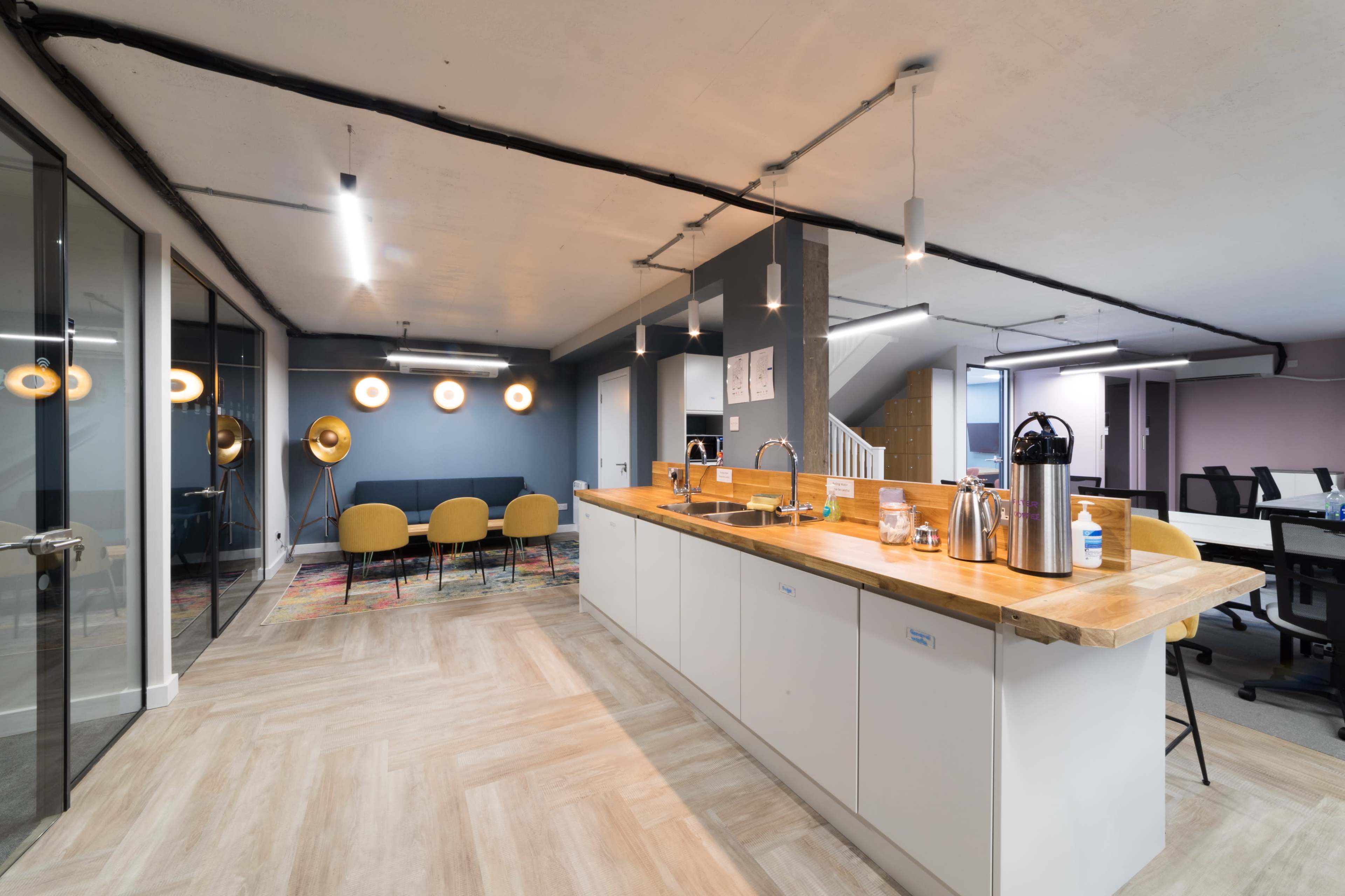 A modern office space featuring a kitchenette with a wooden counter, seating area with yellow chairs, and workstations in the background.