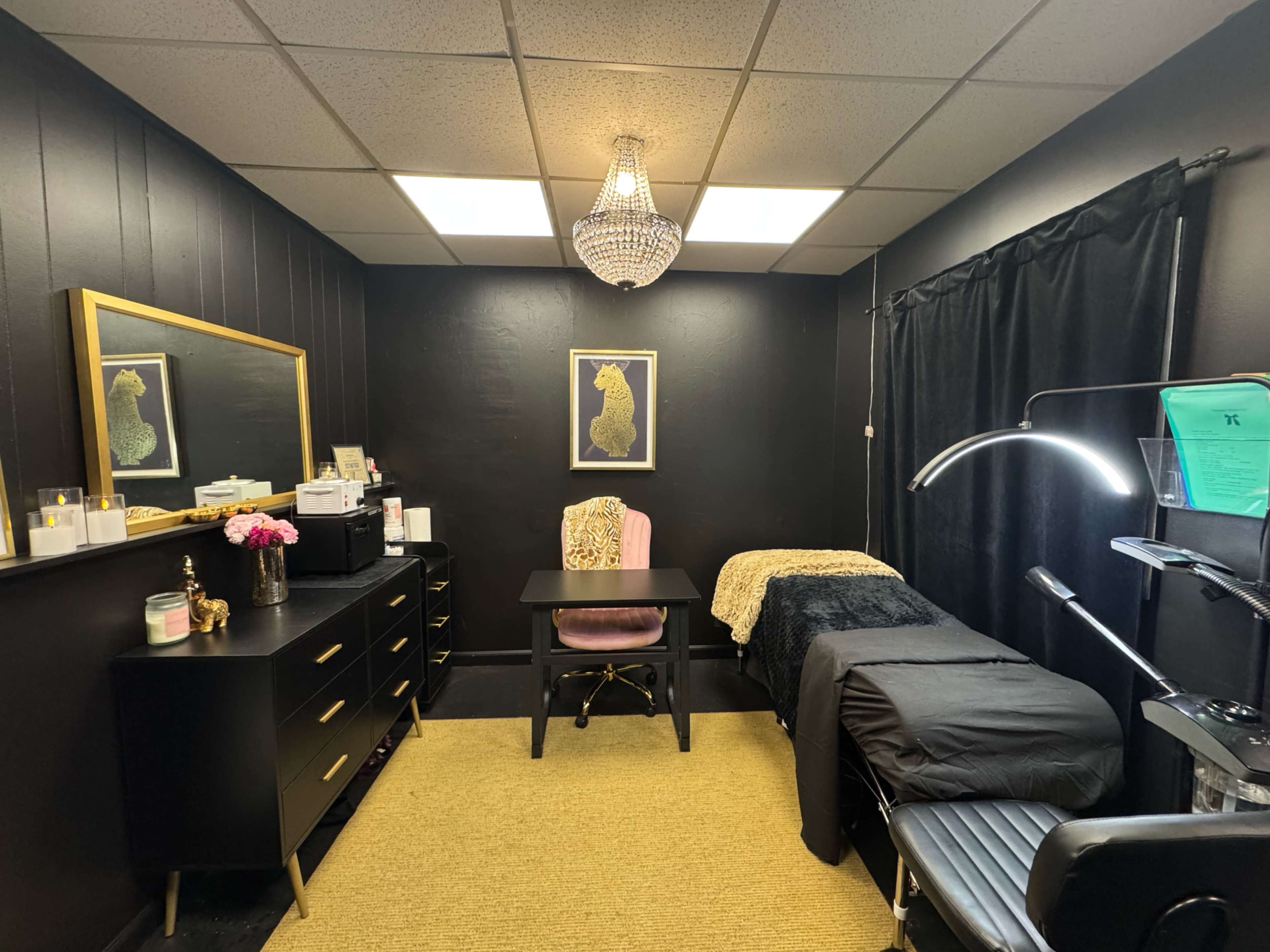 The image shows a small, dimly lit room with black walls, featuring a desk, a massage table covered with a dark blanket, a large mirror with a gold frame, and a chandelier hanging from the ceiling.