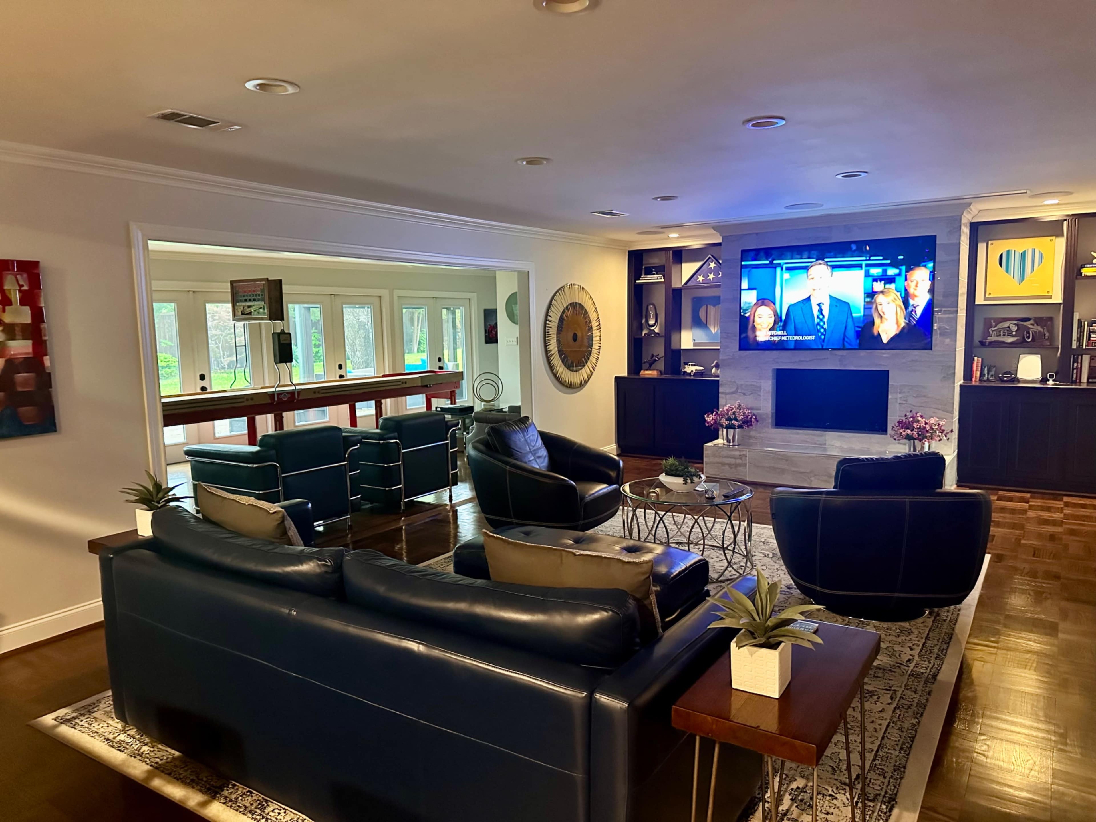 The image shows a modern living room with dark blue sofas, a coffee table, and a large television mounted on a stone wall, overlooking a dining area with bar seating and large windows.