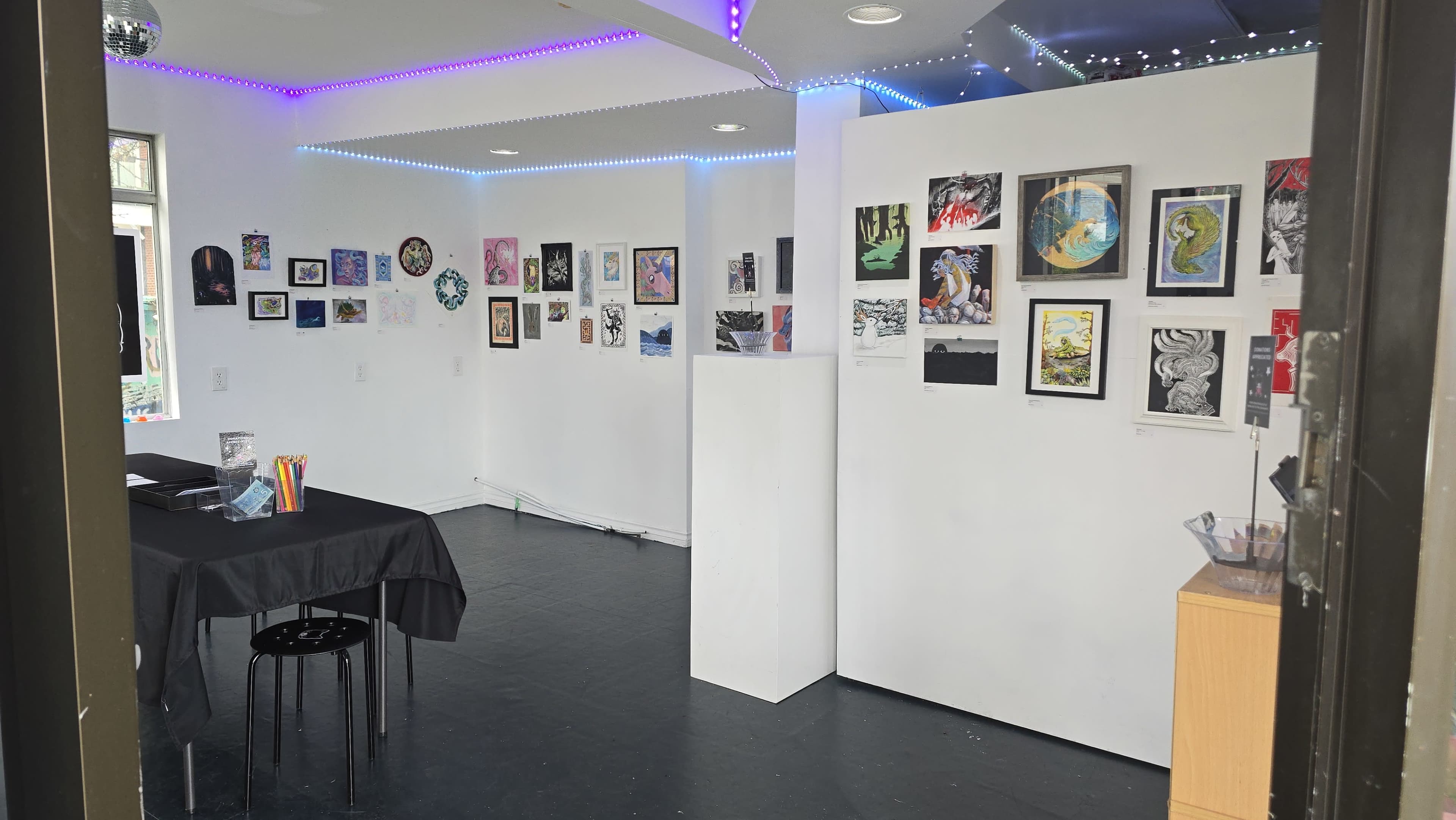 The image shows an art gallery with white walls displaying various framed artworks and colorful lighting decorations.