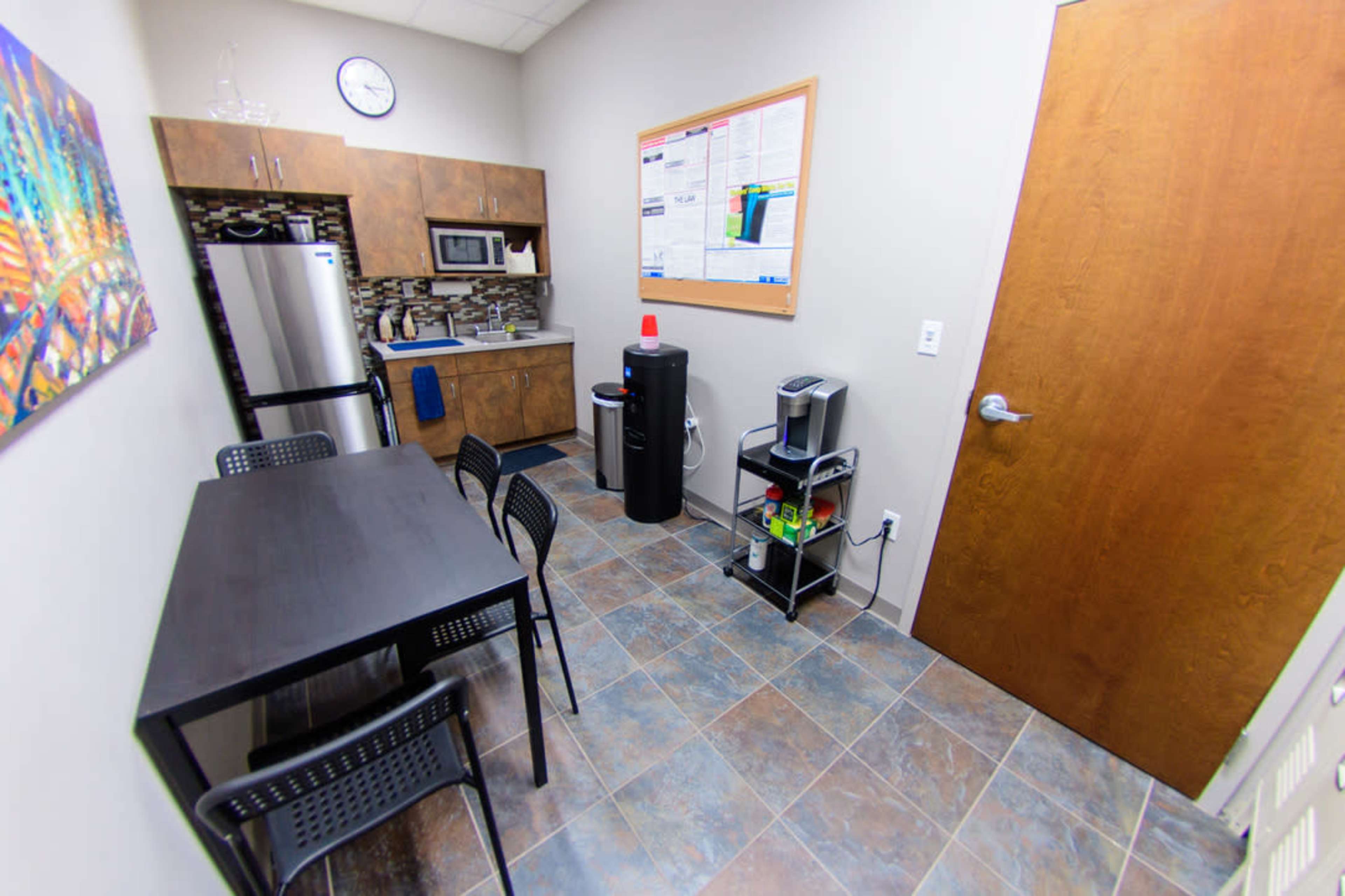 A small kitchen area with a table, chairs, refrigerator, microwave, and a water cooler, located next to a door.