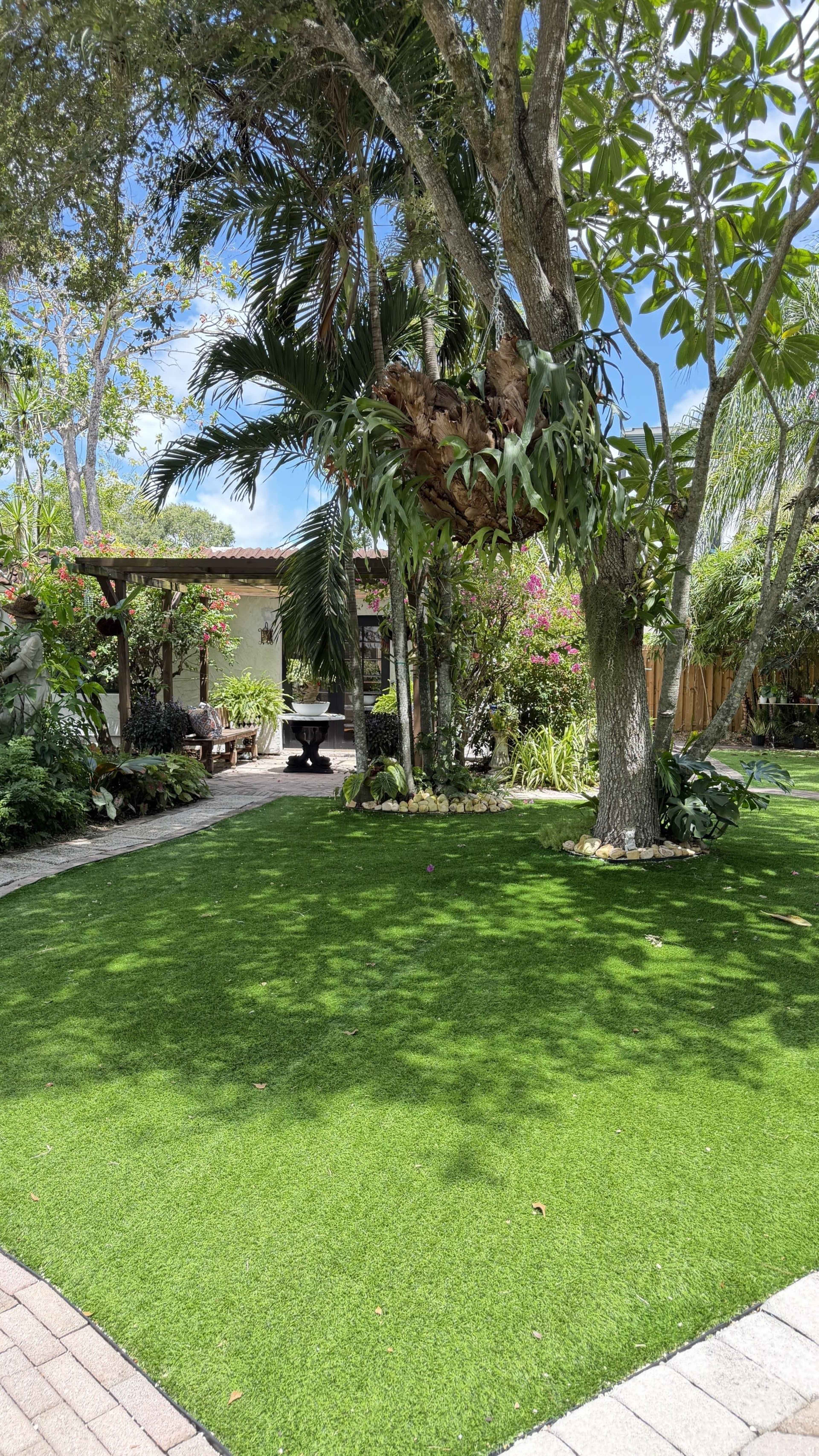 A landscaped yard features artificial grass, surrounded by various trees and shrubs, with a patio area visible in the background.