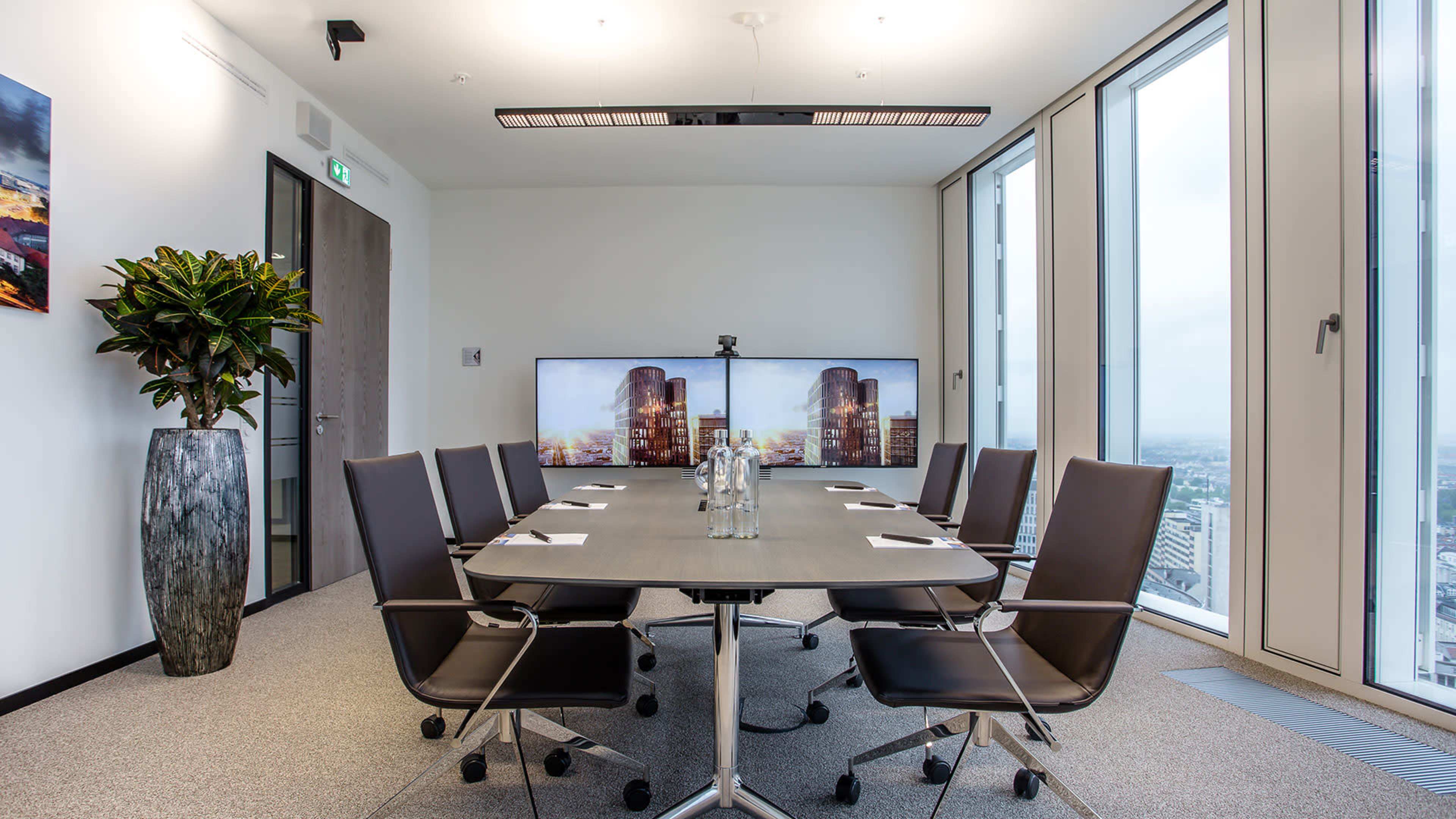A modern conference room with a rectangular table, black chairs, two large screens displaying cityscapes, and a large plant in a corner.