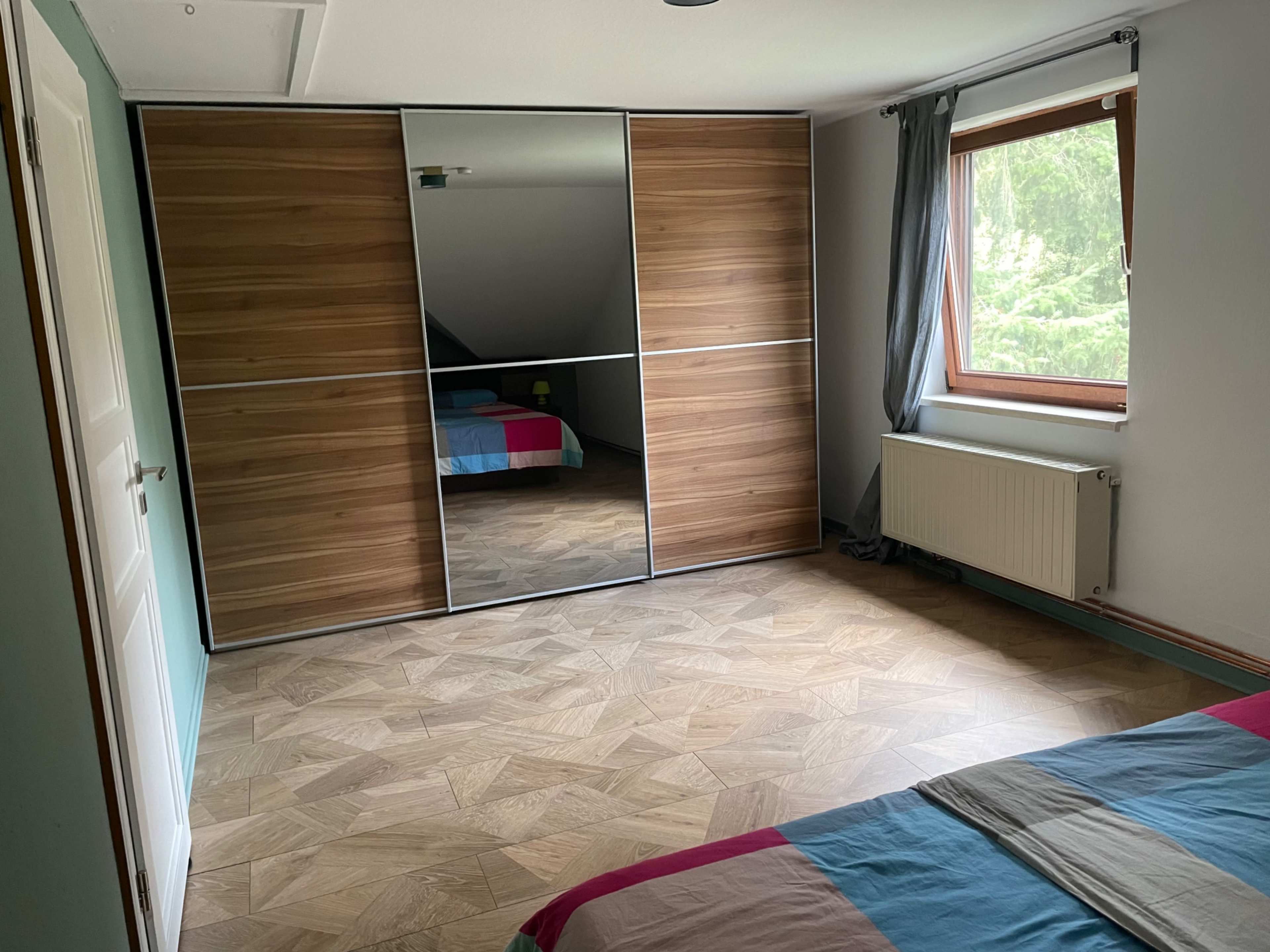 The room features a wooden floor, a large mirrored wardrobe, a bed with colored bedding, and a window overlooking greenery.