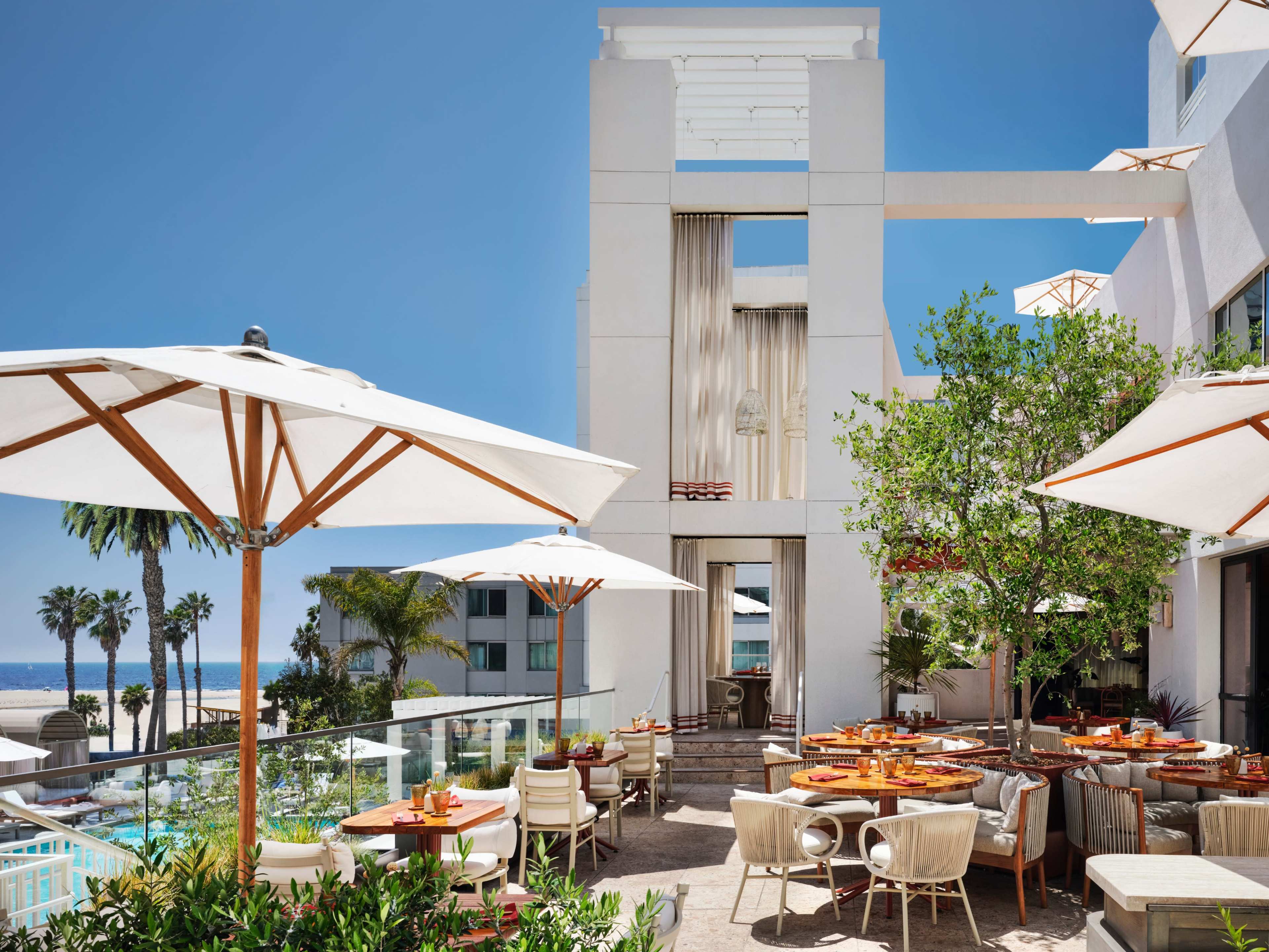 The image depicts a bright outdoor dining area at a modern restaurant, featuring white umbrellas, wooden tables, and a view of palm trees and a pool.