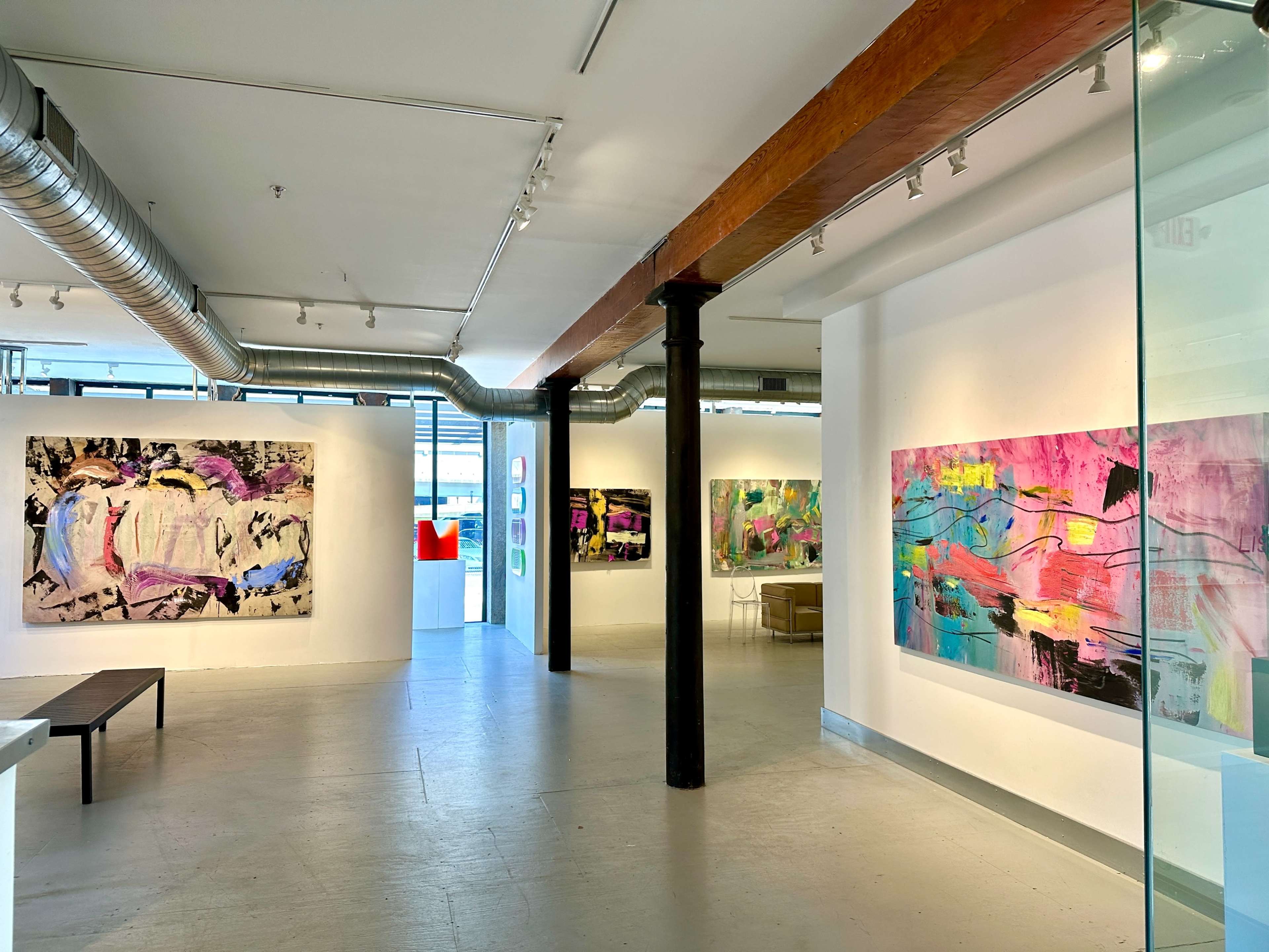 The image shows an art gallery featuring abstract paintings on white walls, with a bench in the foreground and large windows allowing natural light to fill the space.