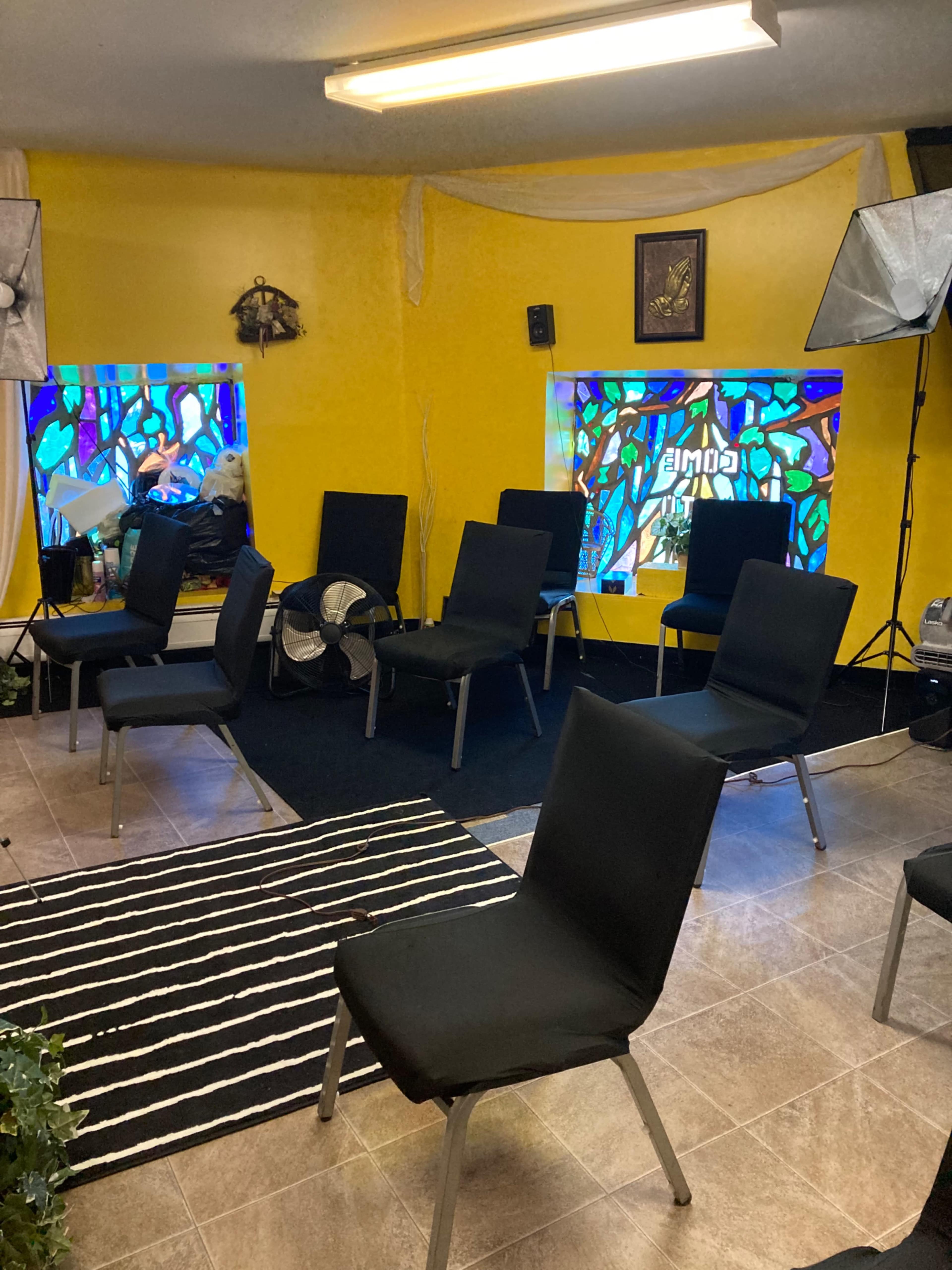 A room with several black chairs arranged in a circle, surrounded by yellow walls and windows featuring stained glass designs.