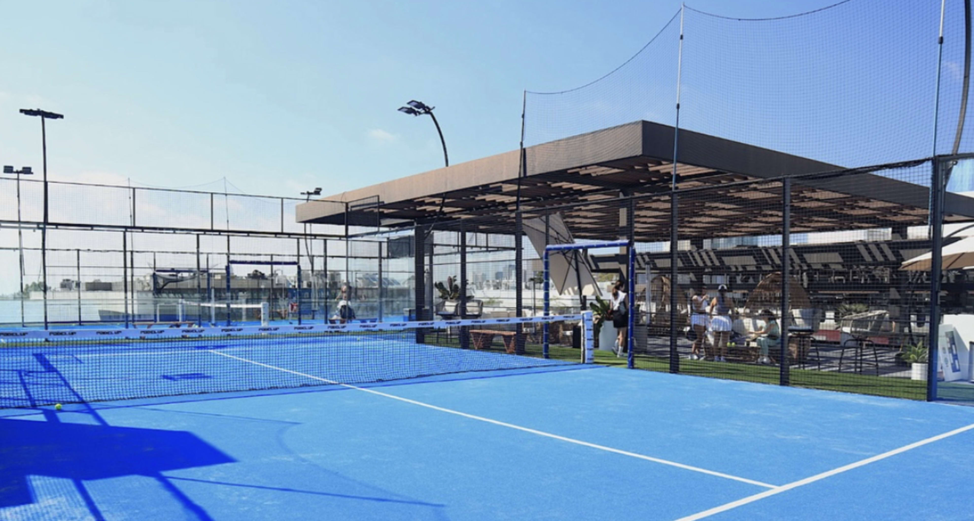 The image shows a blue paddle tennis court next to a modern outdoor seating area with a roof and netting.