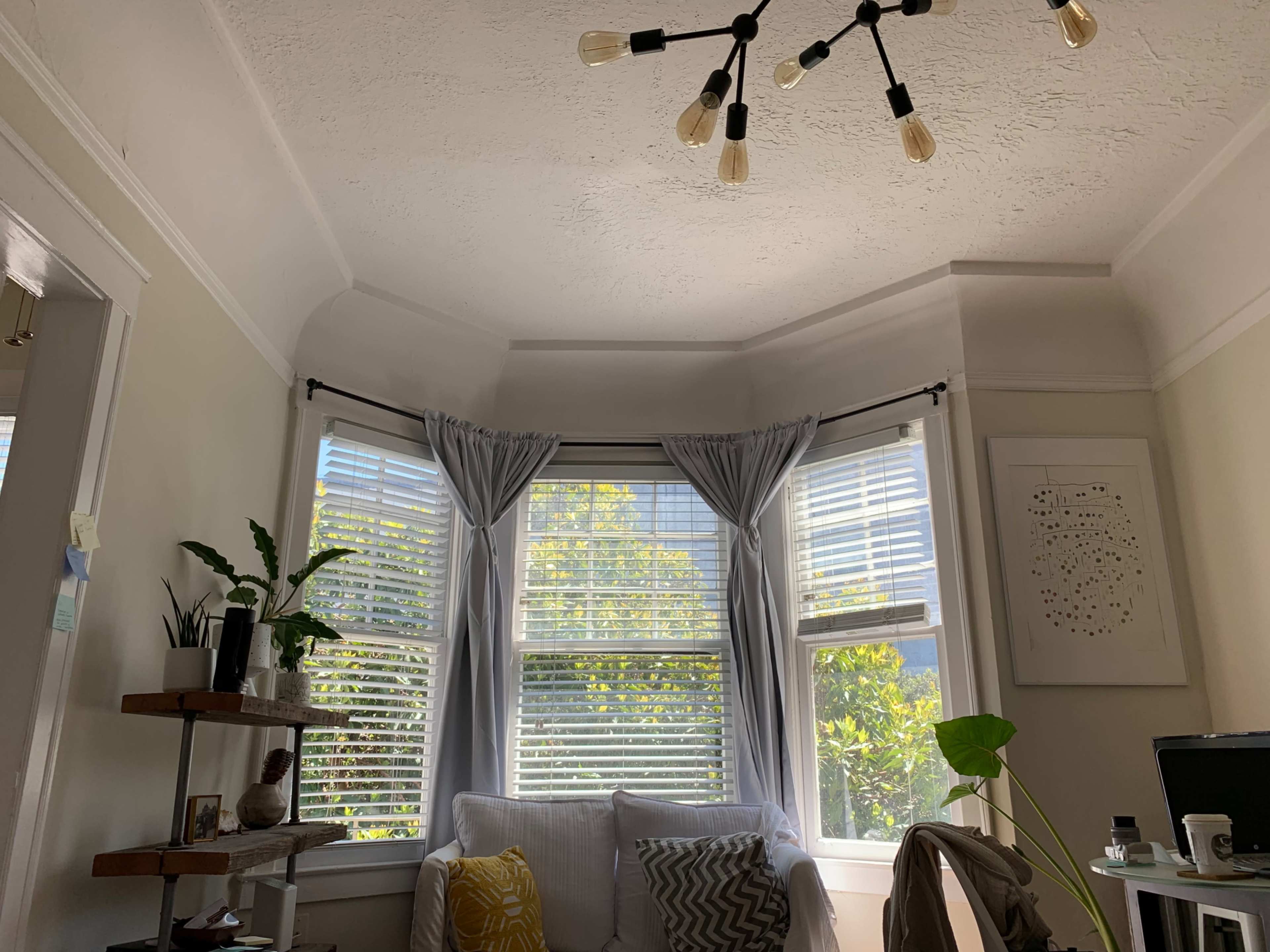 A cozy living room corner with a bay window adorned with light curtains, a small plant on a shelf, and a white sofa.