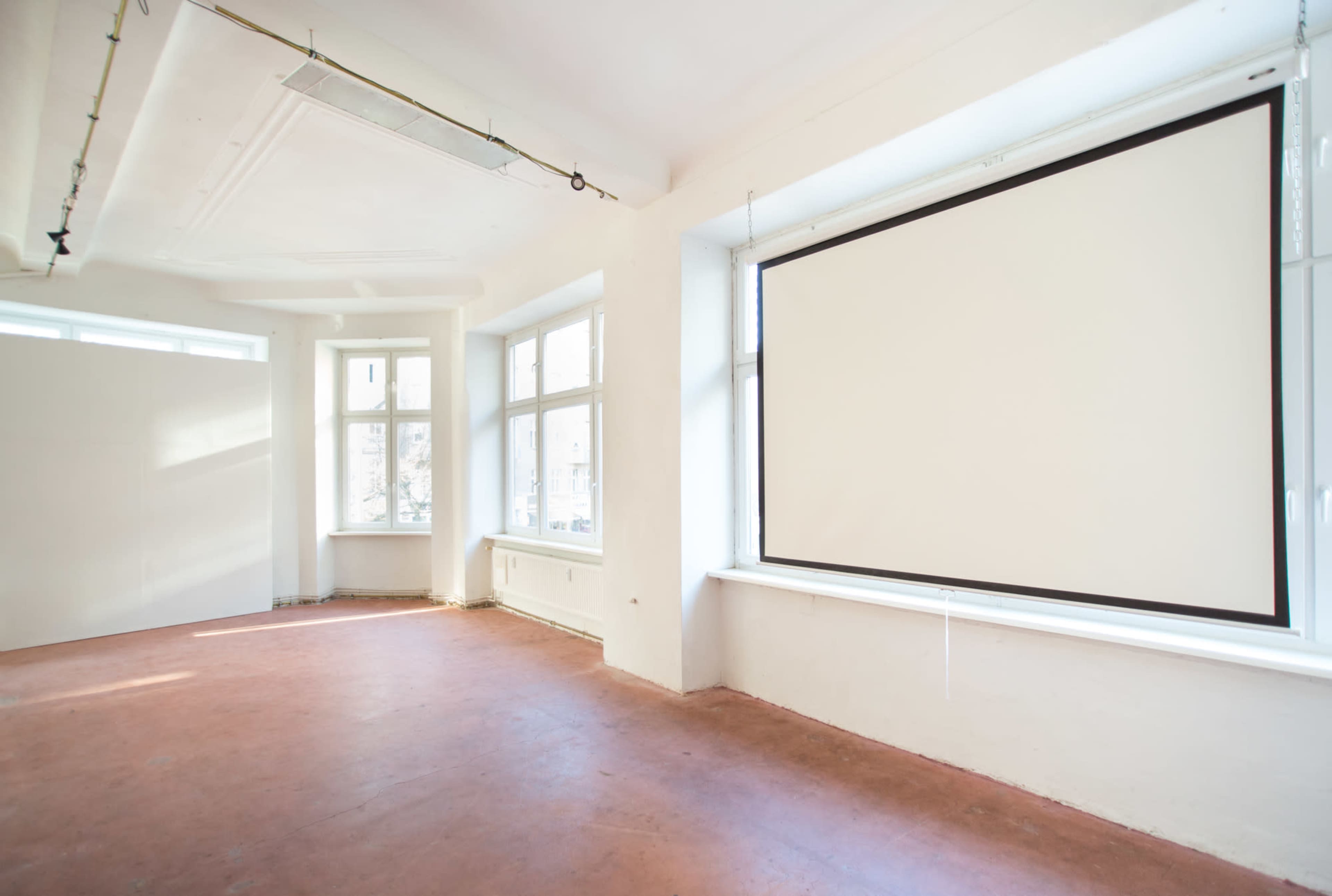 A bright, empty room with large windows and a retractable projection screen mounted on a wall.