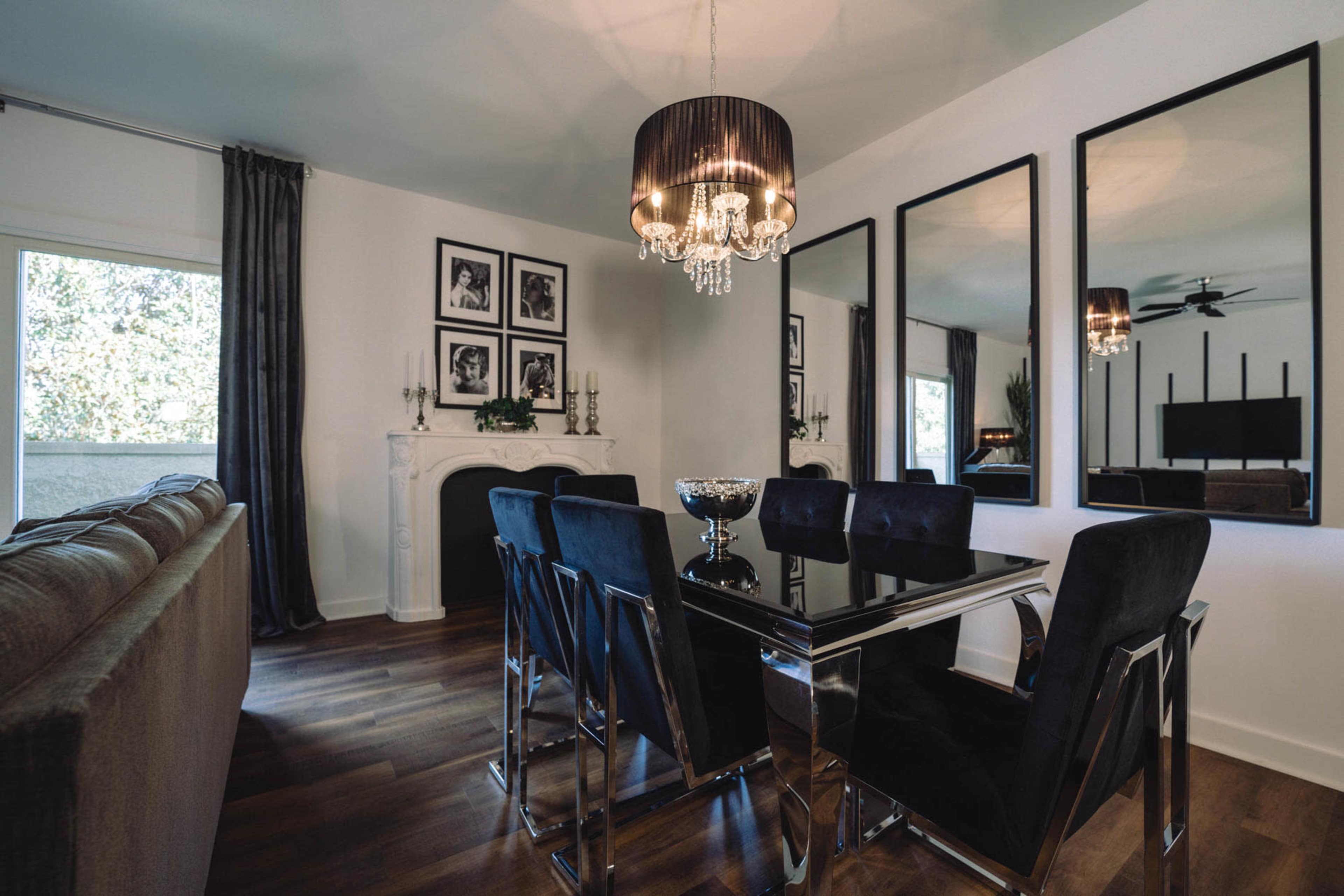 A modern dining area featuring a black glass table surrounded by six black upholstered chairs, with a chandelier overhead and framed photographs on the wall.