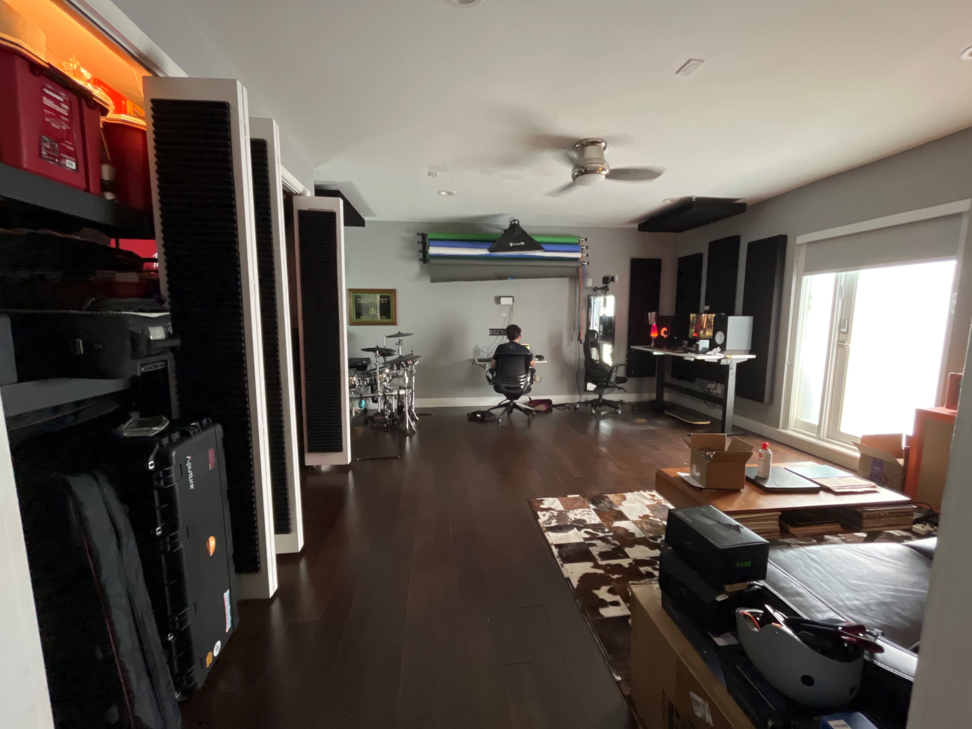 A person sits at a desk in a well-organized room featuring musical equipment, shelving, and a large window.