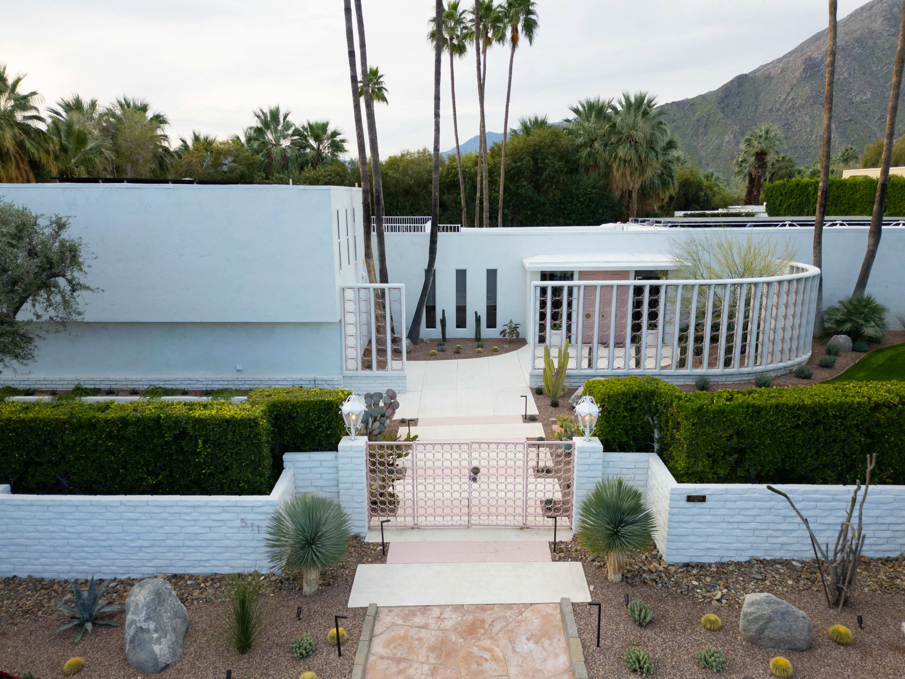 The image shows a modern house with a minimalistic design, surrounded by palm trees and landscaped gardens, featuring a gated entrance and a circular patio area.