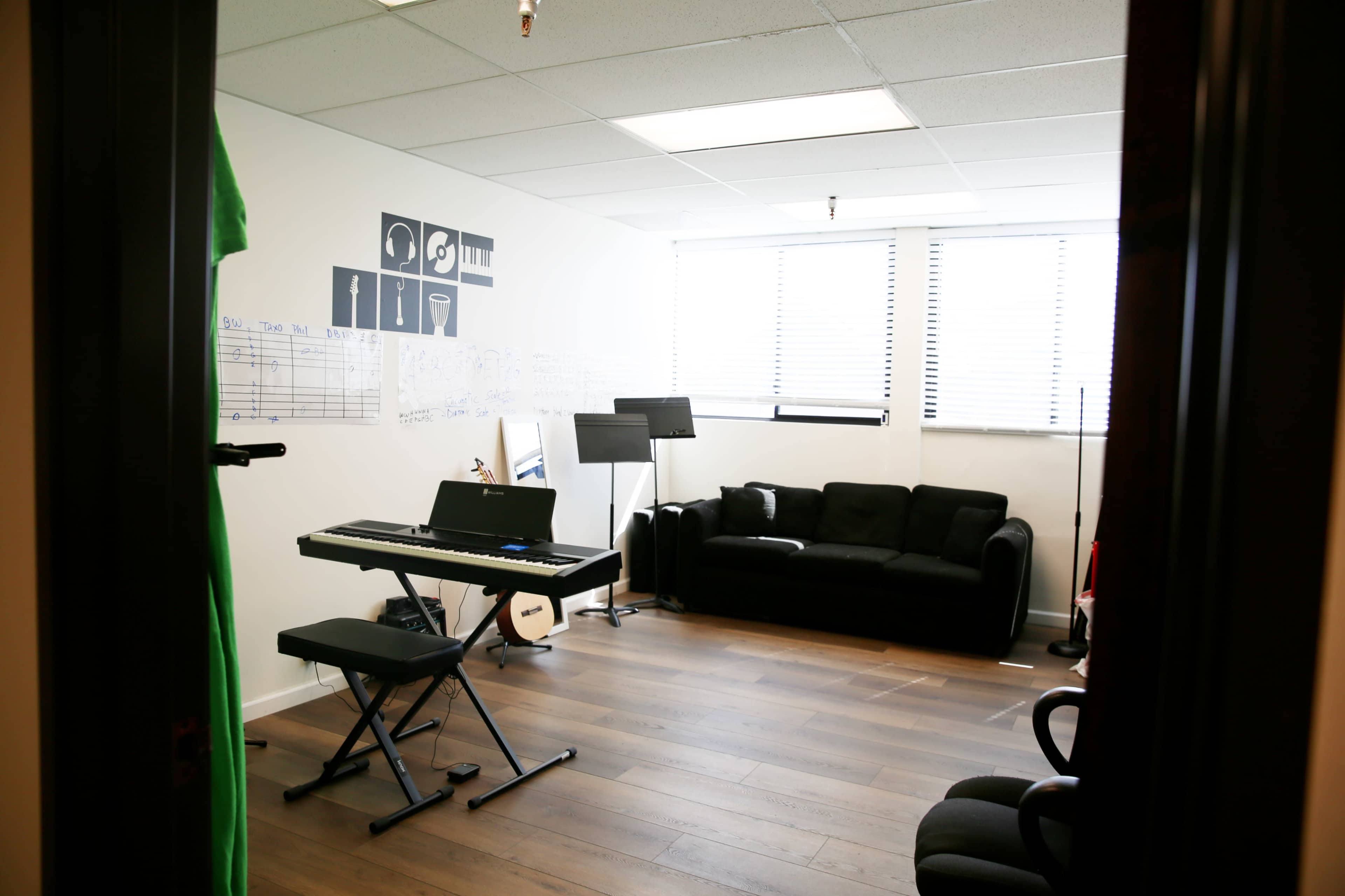 The image shows a music practice room featuring a keyboard, a black couch, sheet music on display, and a music stand.