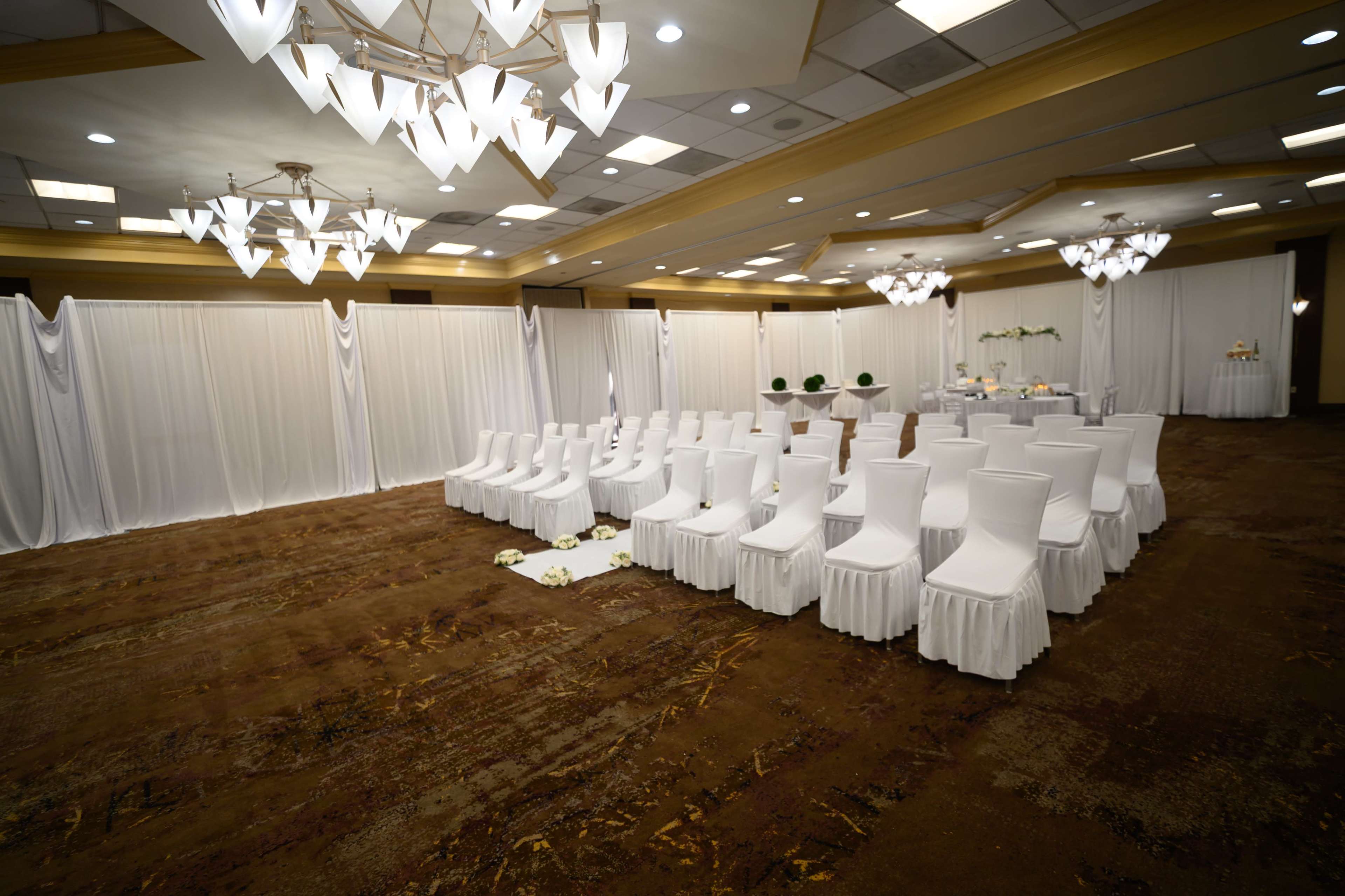 The image shows a conference room set up for a wedding ceremony, featuring white chairs arranged neatly in rows and elegant lighting.