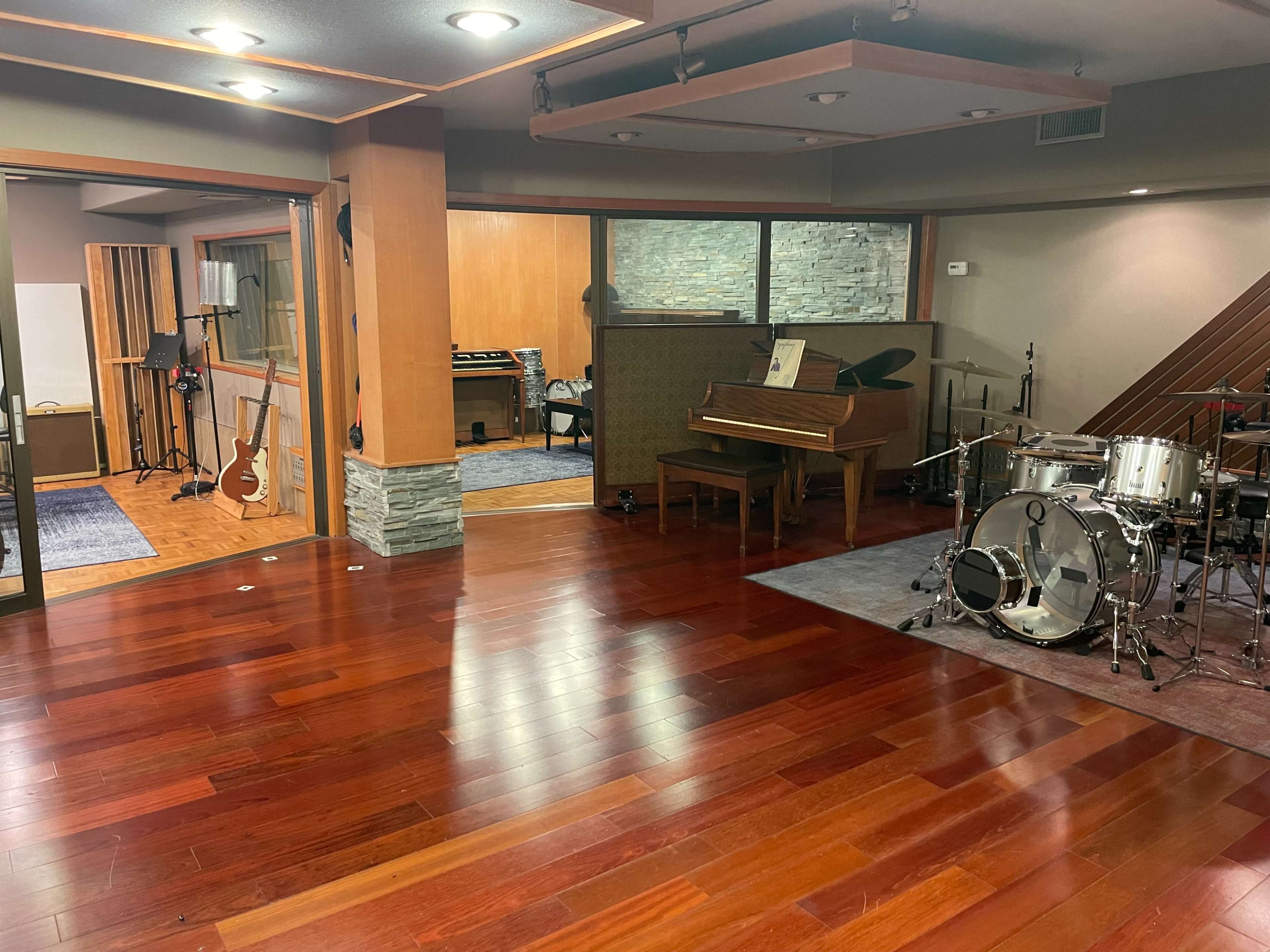 A music studio features a wooden floor, a grand piano, a drum set, and various recording equipment visible in the background.