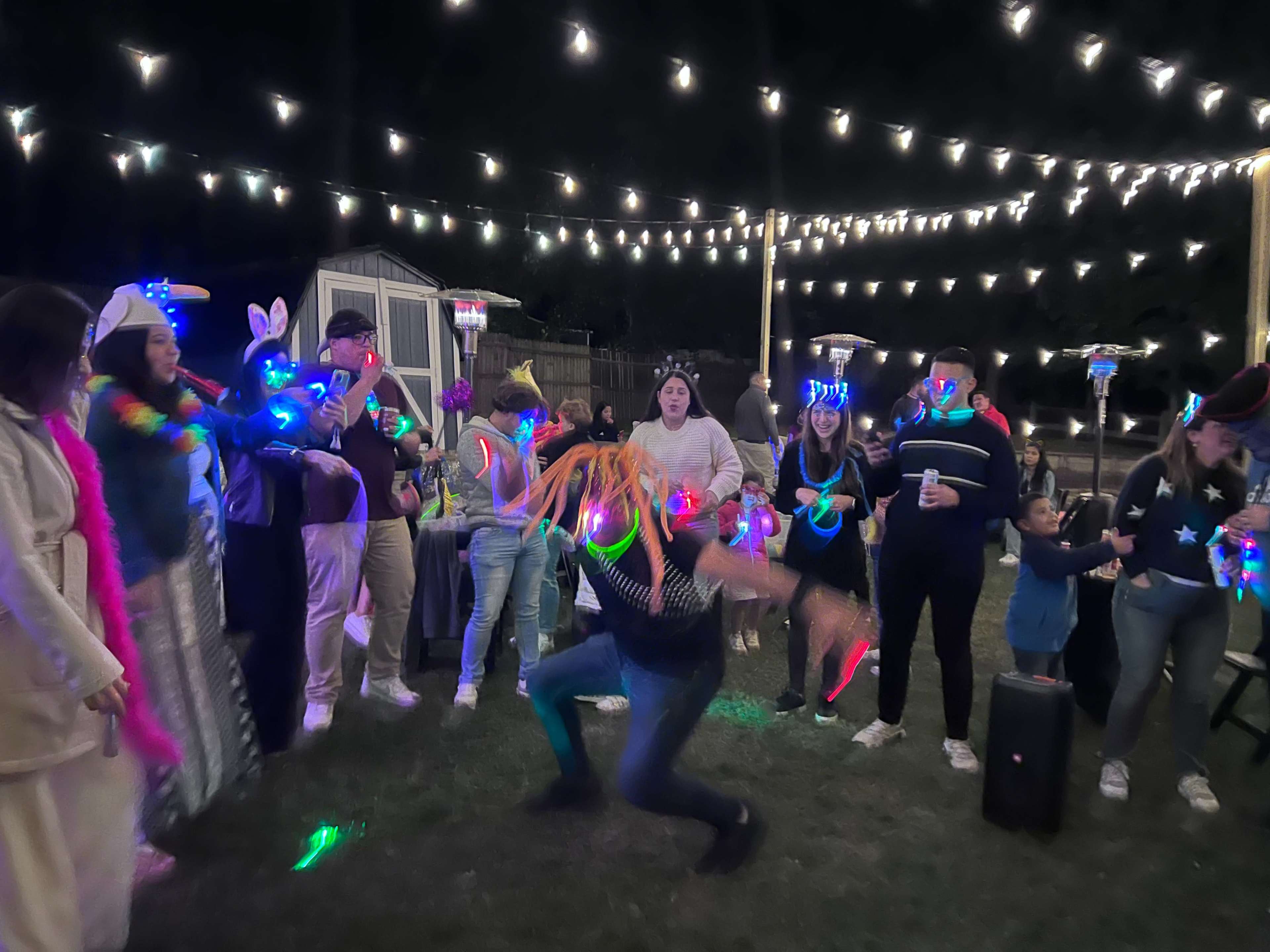 A group of people in festive attire dance and celebrate under string lights at an outdoor party.
