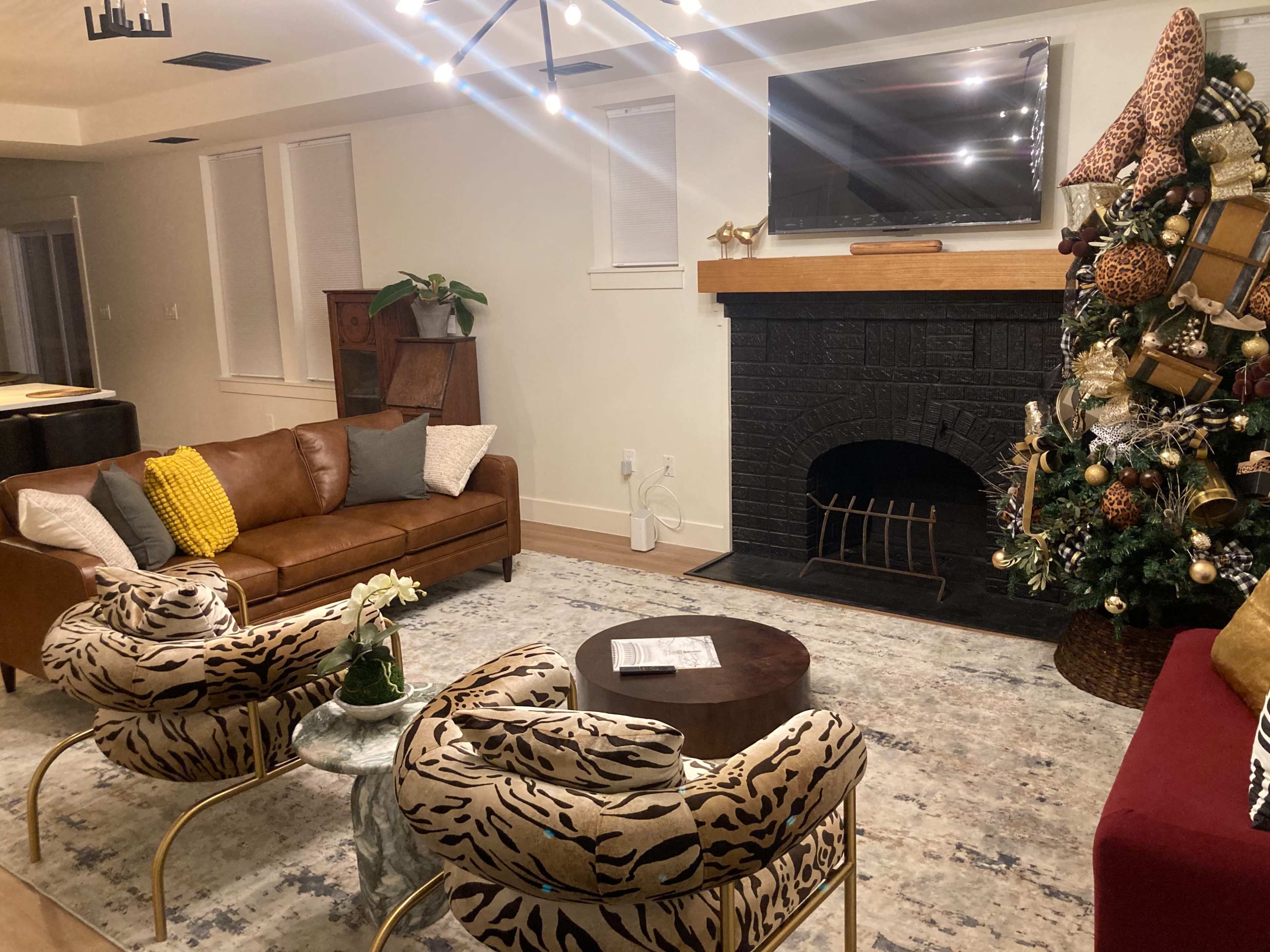 A living room features a brown leather sofa, two animal-print chairs, a coffee table, and a decorated Christmas tree beside a black fireplace with a wooden mantel.