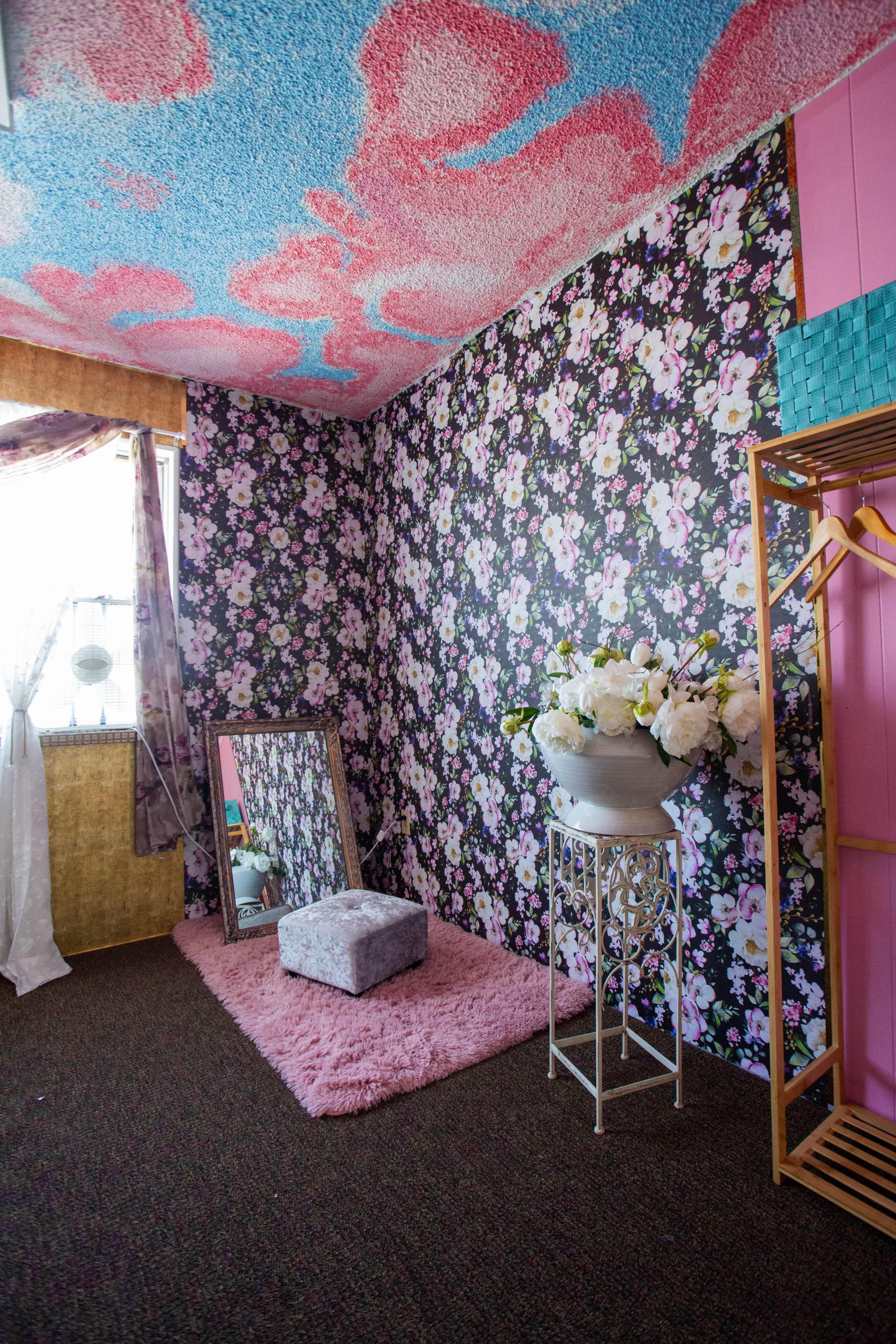 The room features floral wallpaper, a mirrored vanity, a small gray stool, and a decorative flower arrangement, with a distinctly patterned ceiling.