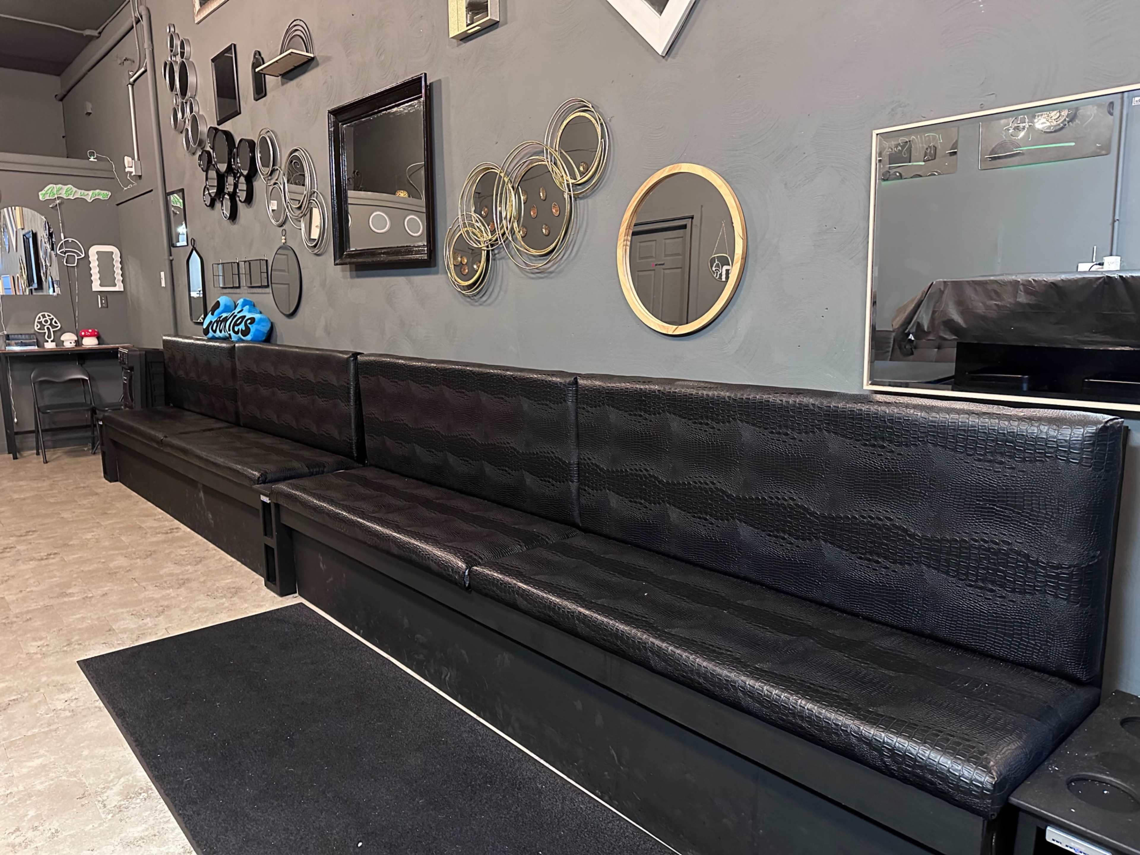 The image shows a long black bench with textured upholstery against a gray wall adorned with various mirrors and circular decorations.