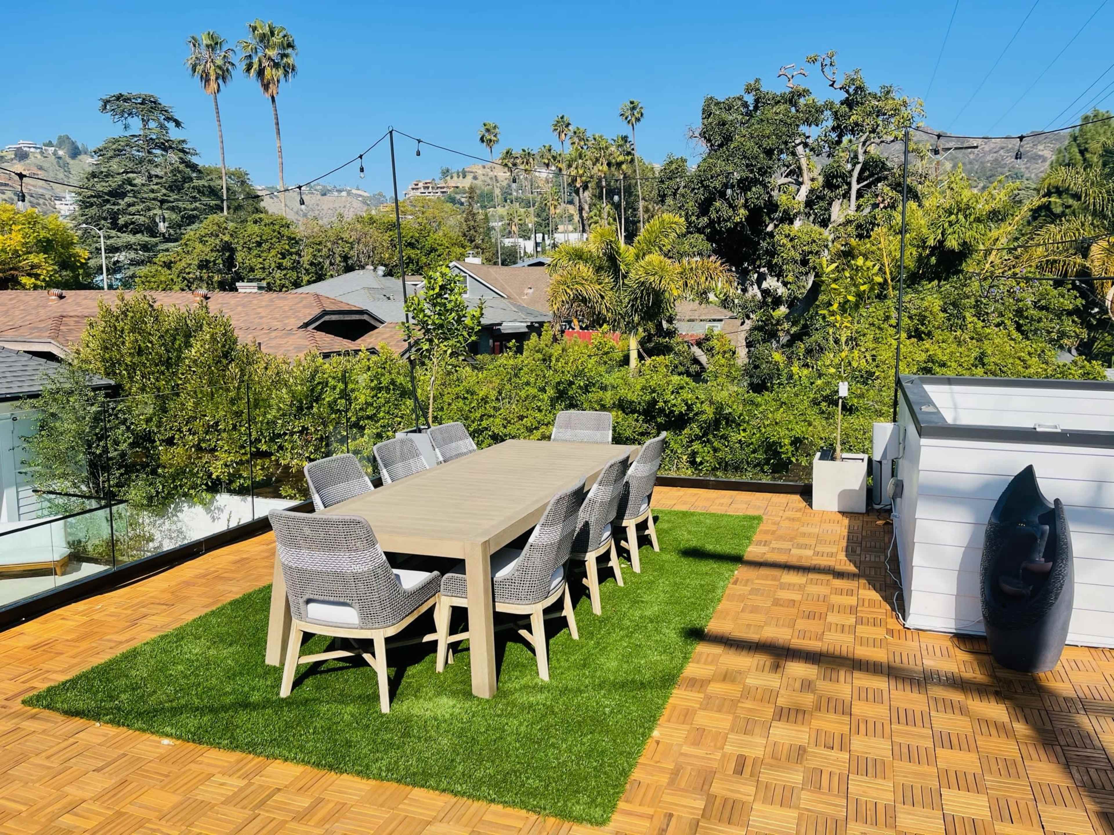 A long dining table with chairs is set on a wooden deck surrounded by greenery and palm trees.