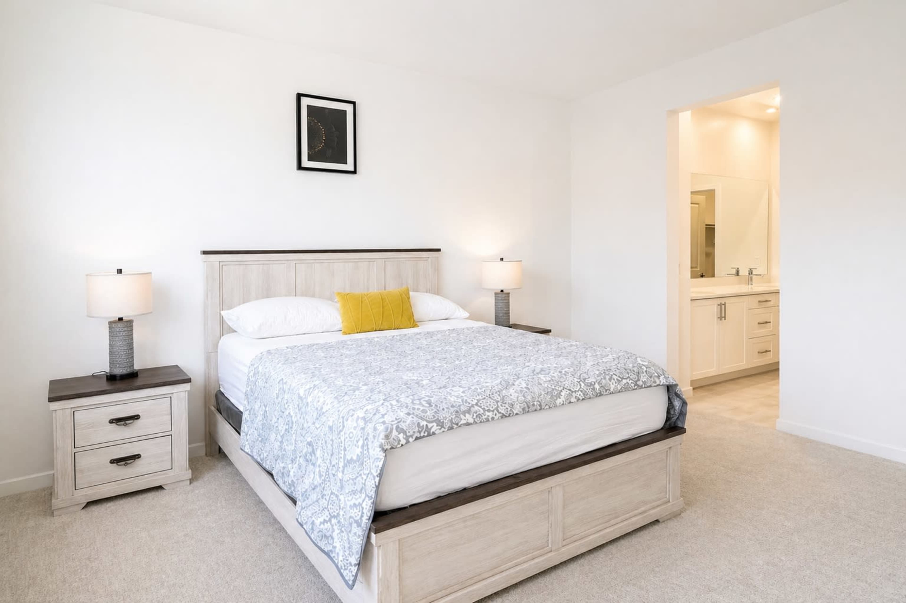 A neatly arranged bedroom features a light wood bed with gray bedding, flanked by two lamps and connected to an open bathroom area.
