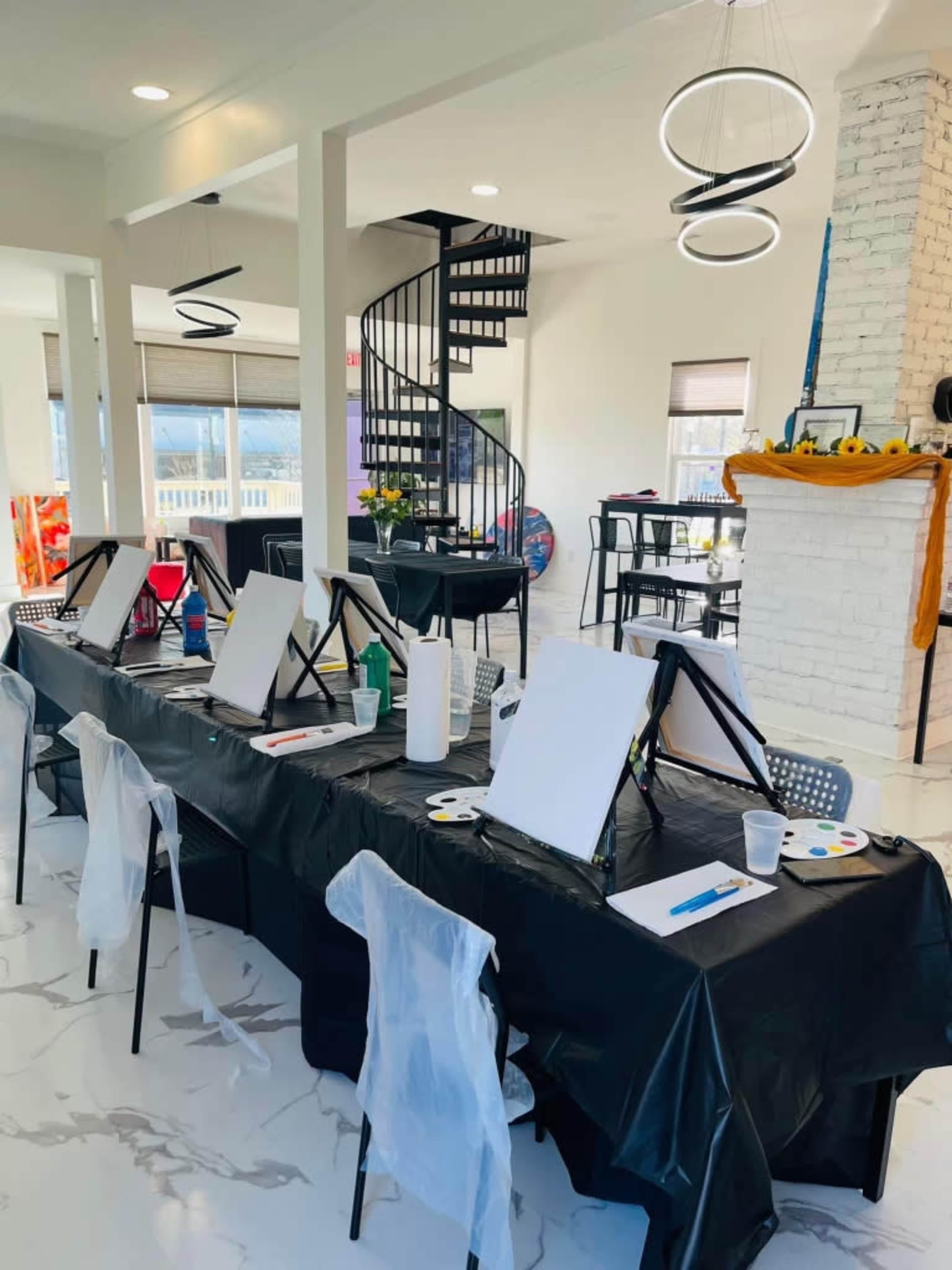 An art studio setup with easels and blank canvases arranged on a long table, while a spiral staircase is visible in the background.