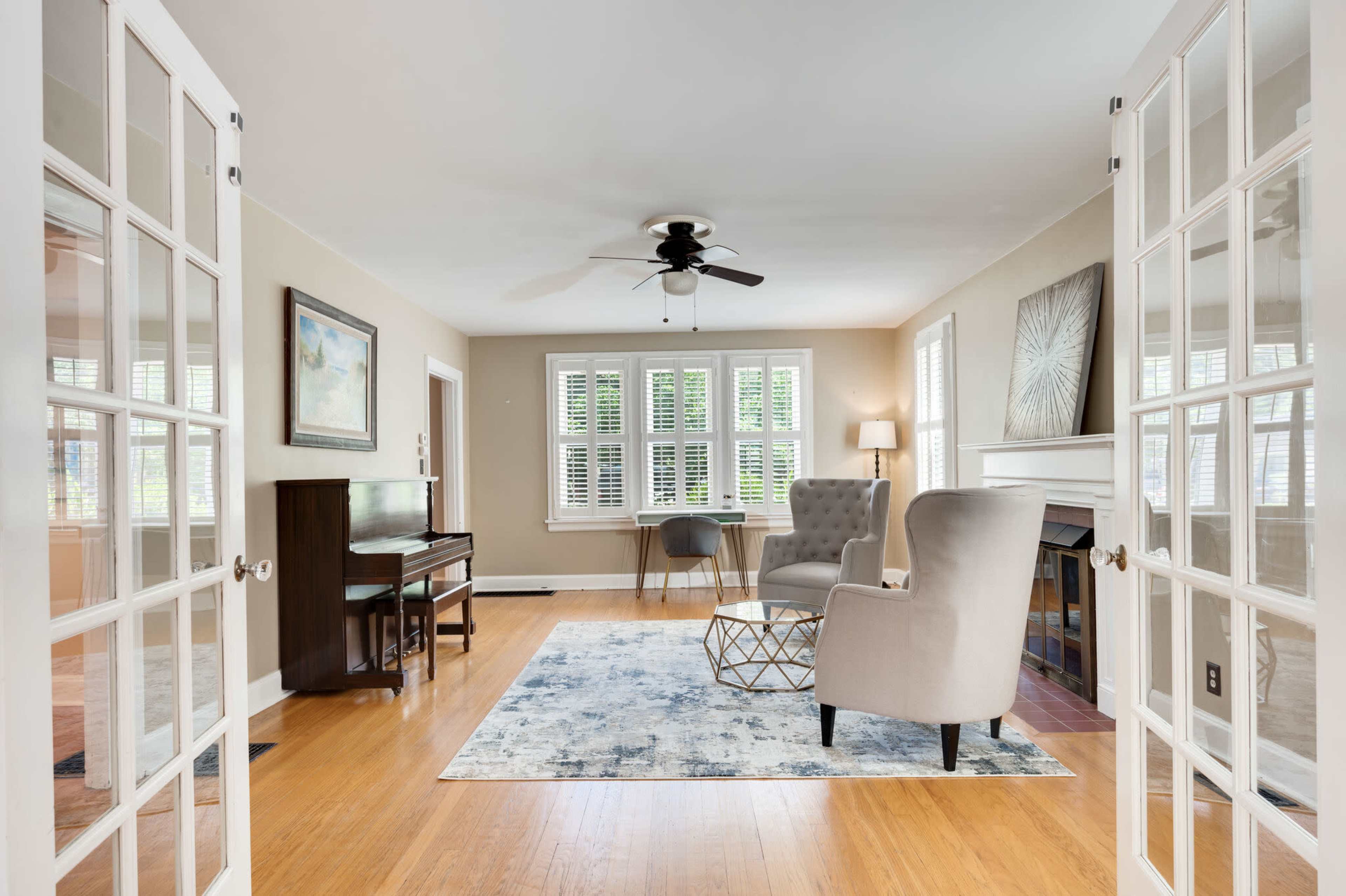 A bright living room features a piano, two armchairs, and a decorative rug, all accentuated by large windows and glass-paneled doors.