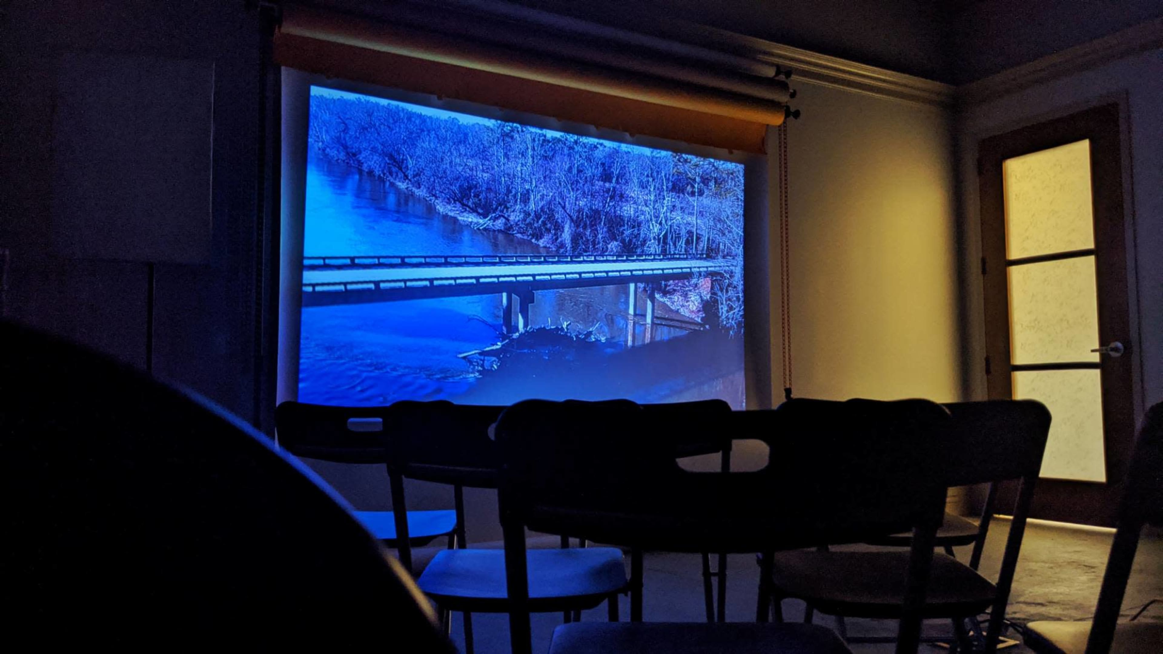 A projection of a river and bridge scene is displayed on a screen in a dimly lit room with empty chairs arranged in front of it.