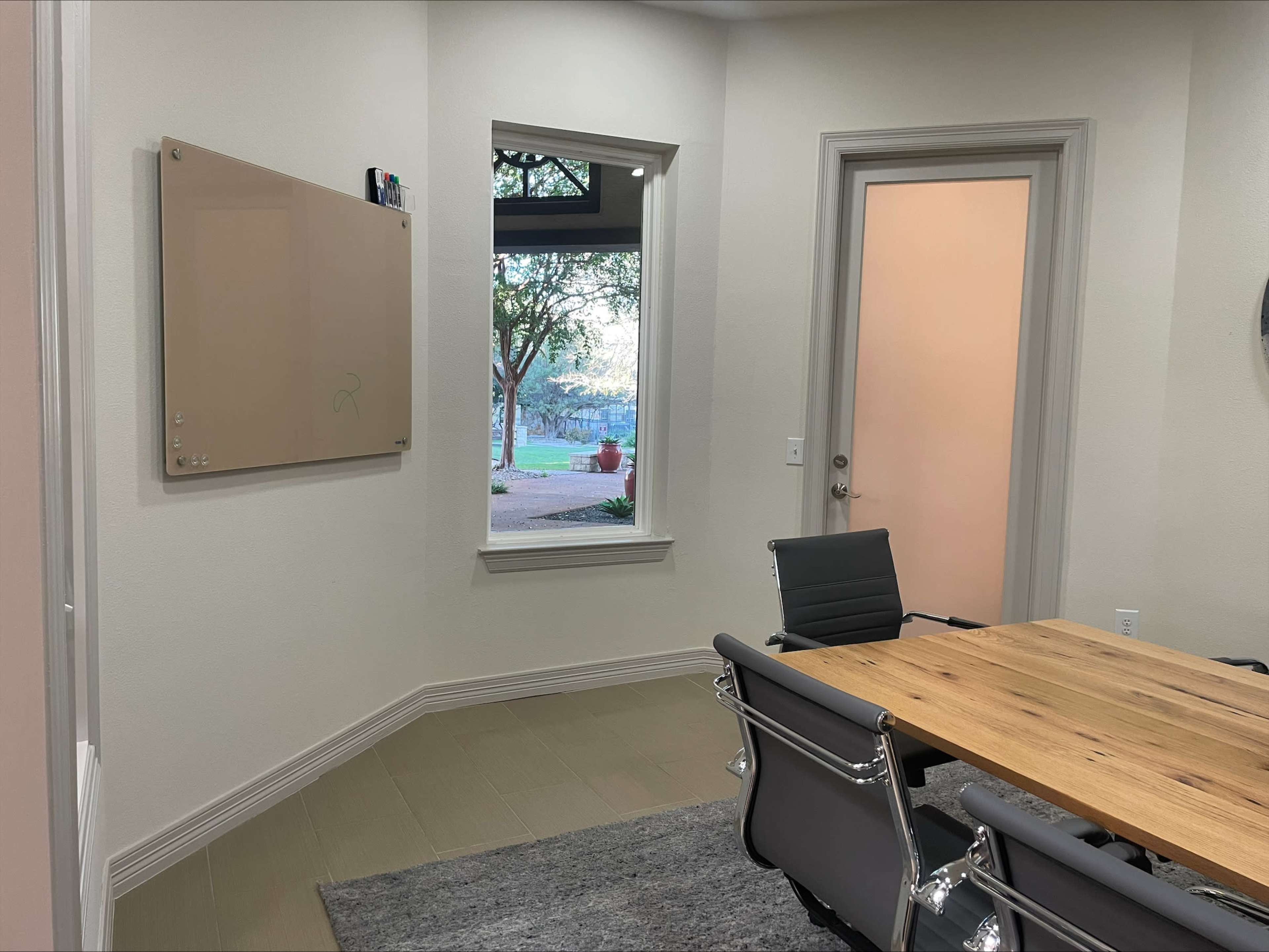 The image shows a modern conference room with a wooden table, ergonomic chairs, a dry-erase board on the wall, and a window displaying greenery outside.