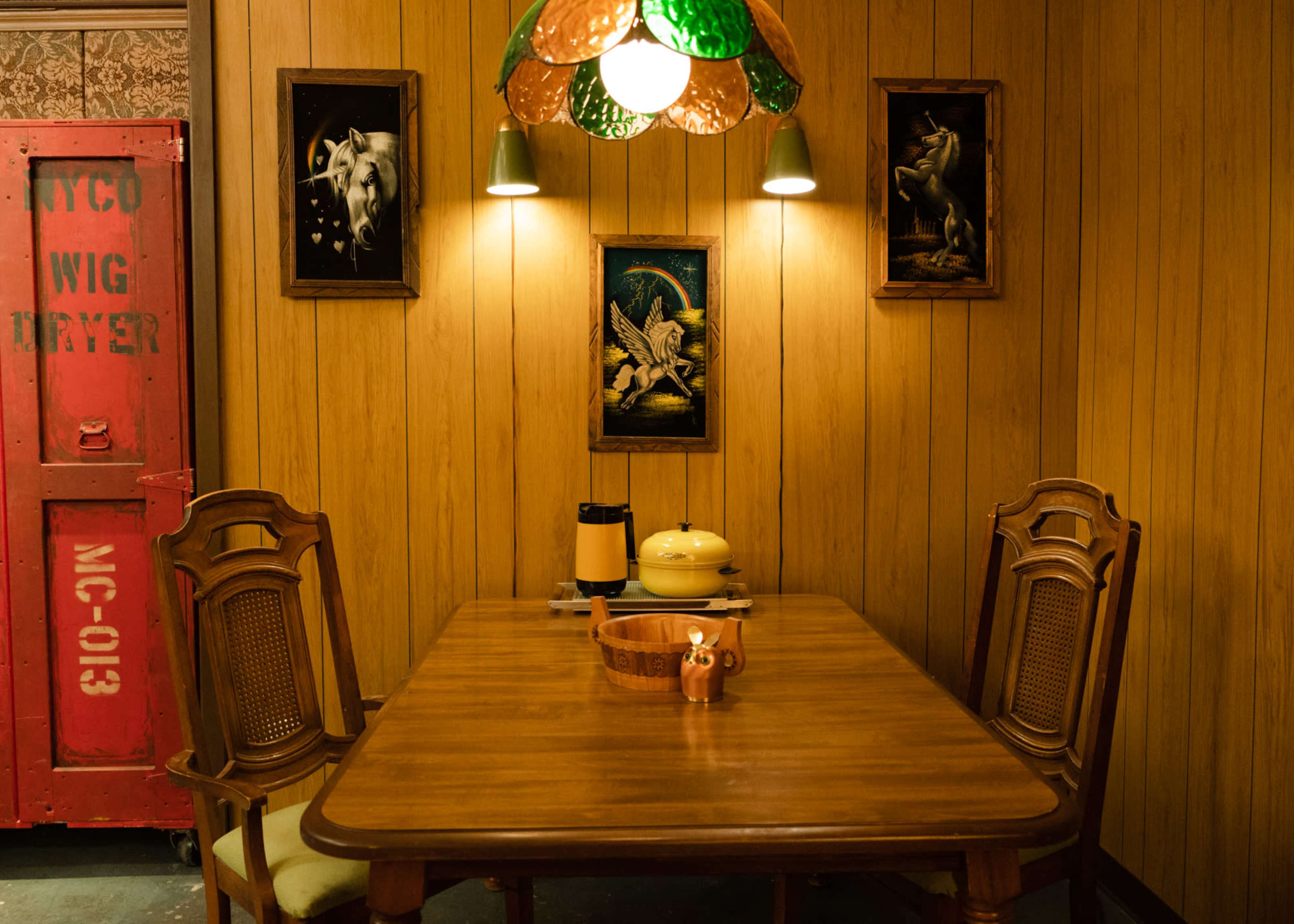 A wooden dining table is set in a room with paneled walls, featuring a vintage lamp, a coffee pot, and framed artwork of animals.
