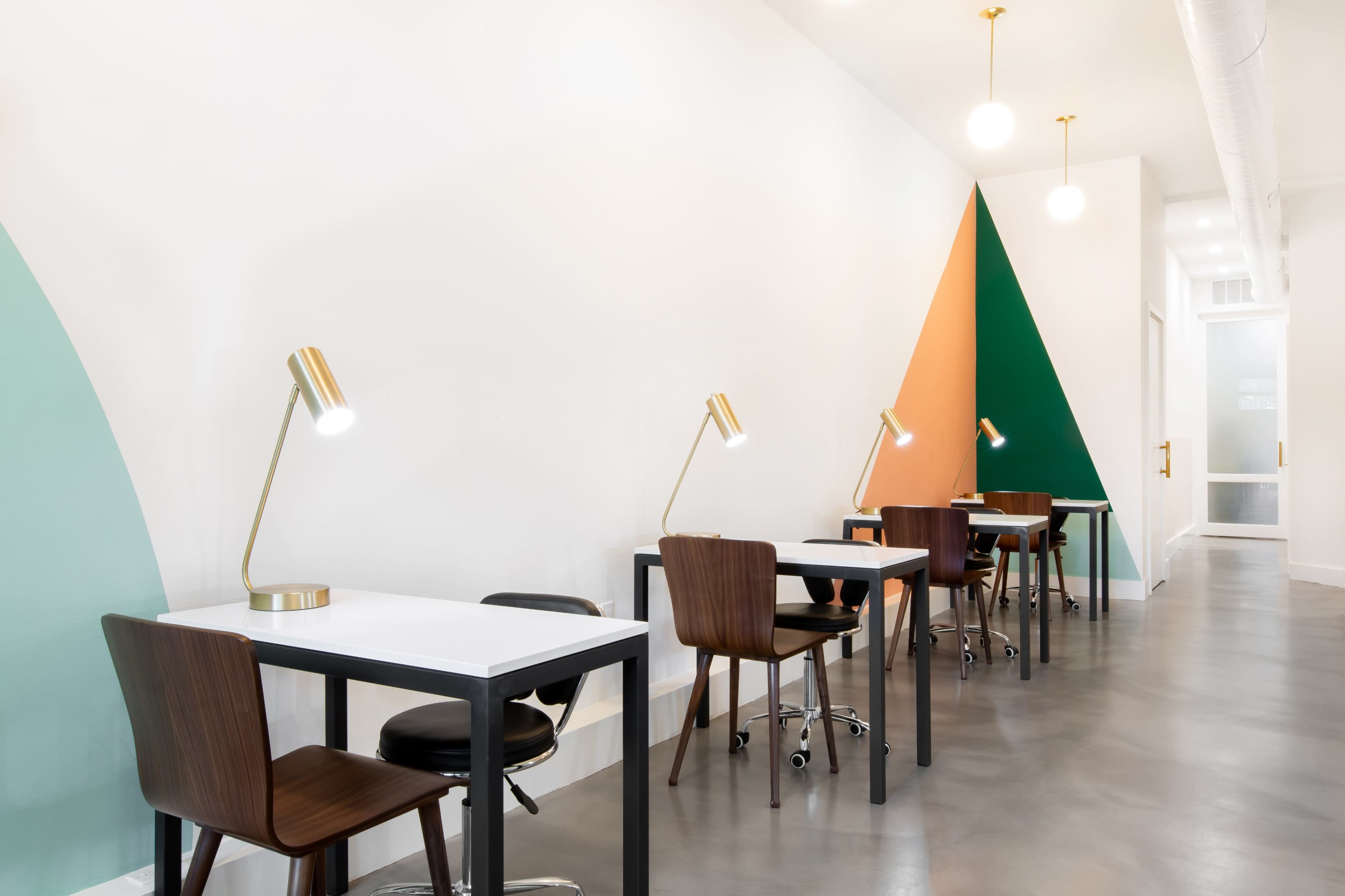 A modern workspace with minimalist tables and chairs, illuminated by stylish desk lamps against a backdrop of colored wall sections.