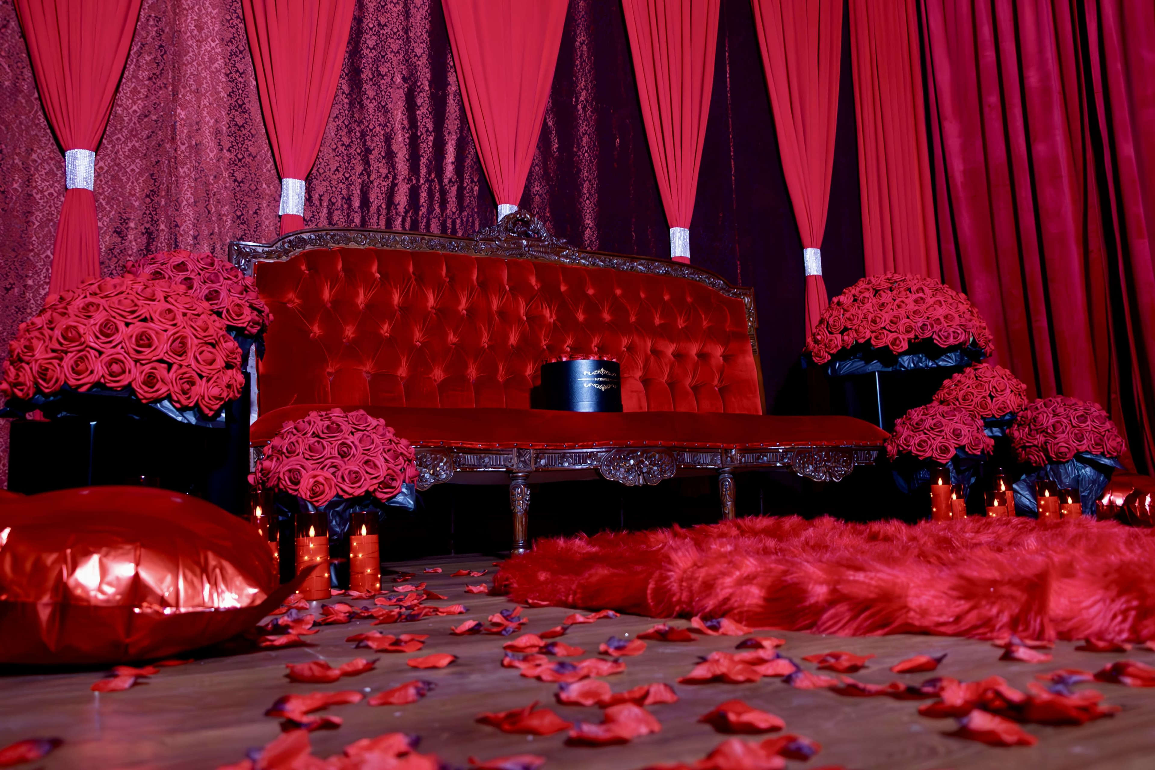 The scene features a richly decorated area with a red velvet sofa surrounded by large bouquets of red roses, red cushions, and scattered rose petals on the floor.