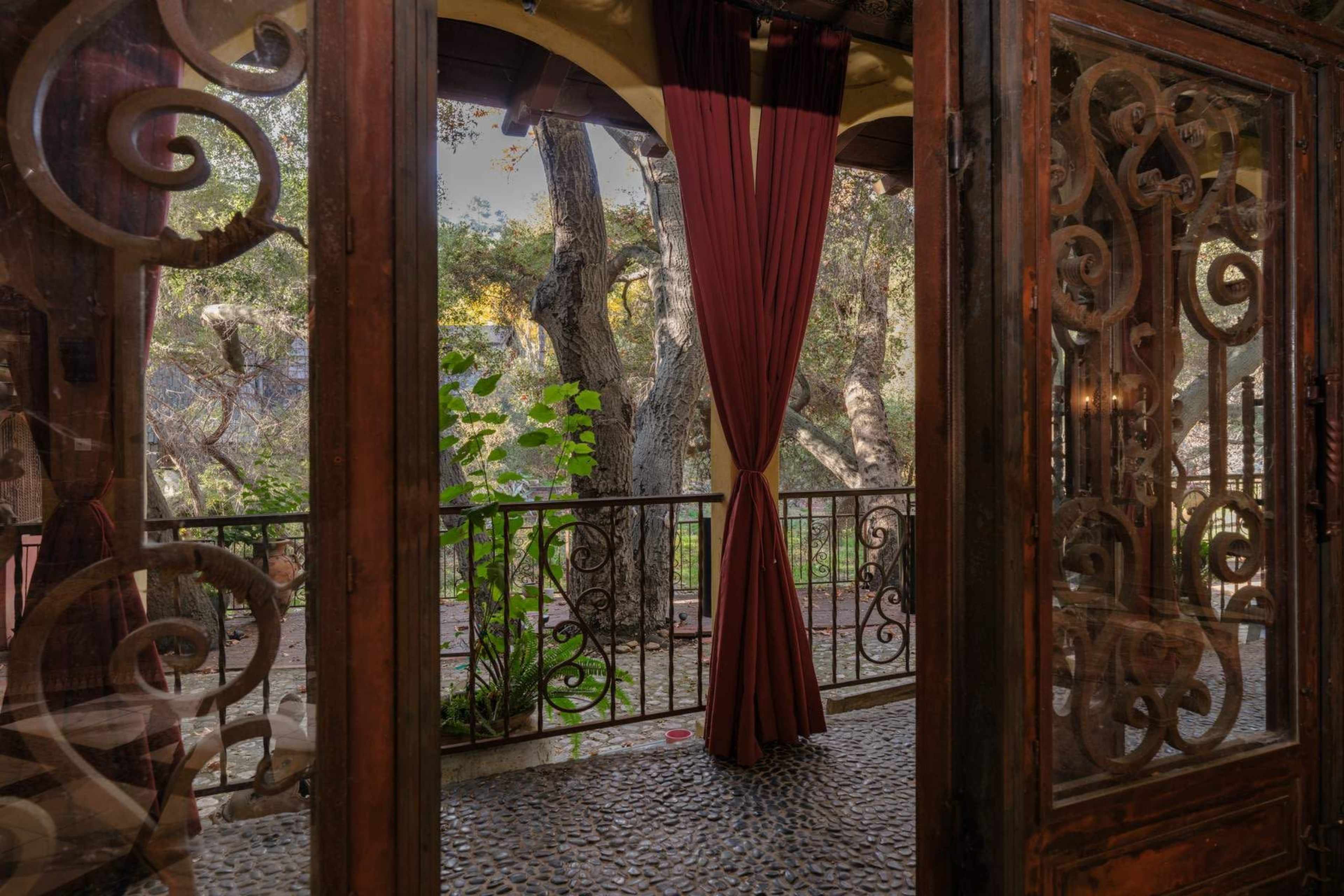 The image shows a wooden-framed balcony with intricate glass designs, overlooking a garden filled with trees.