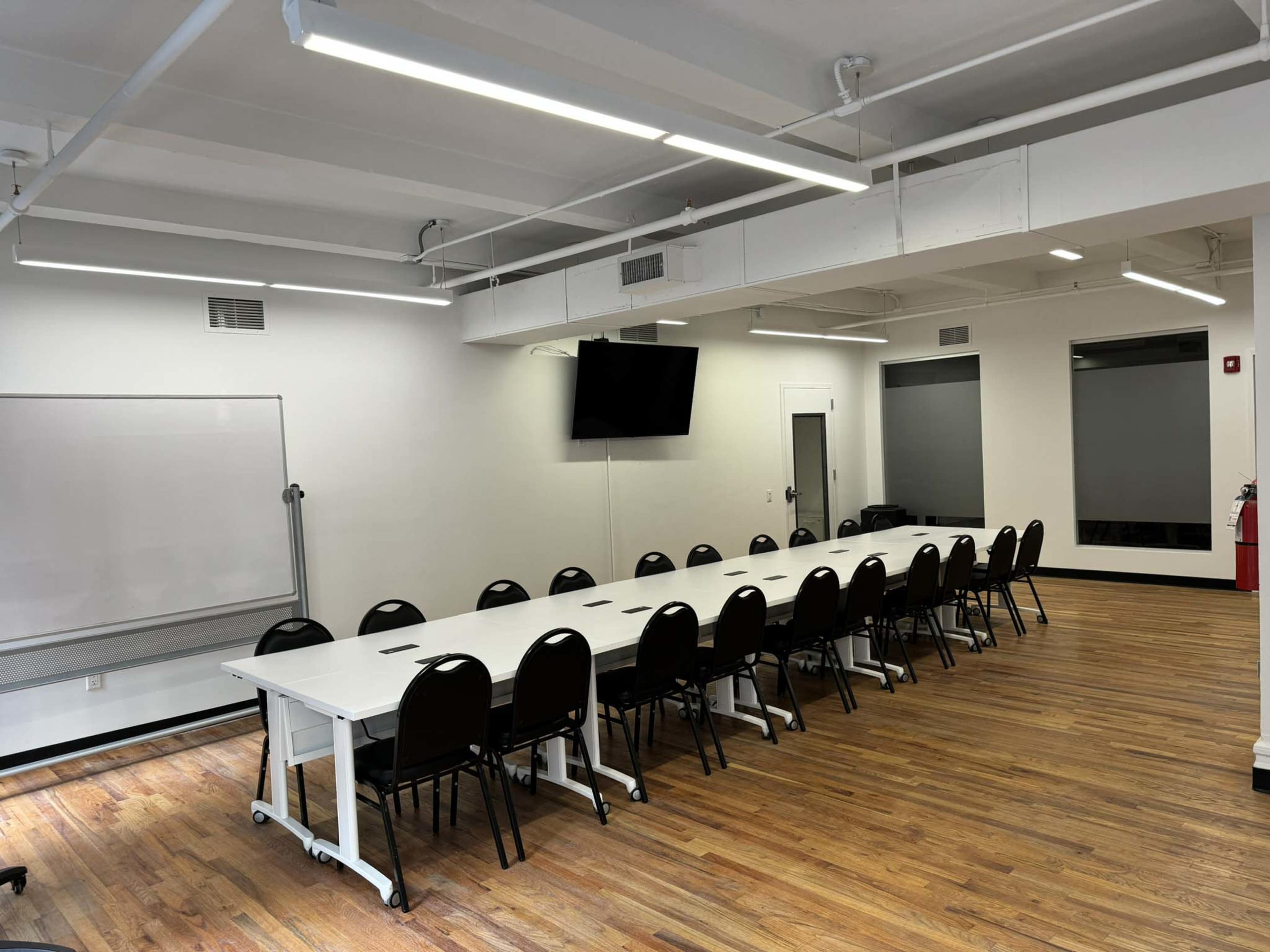A spacious meeting room with a long table surrounded by black chairs, a large monitor on the wall, and whiteboard on one side.