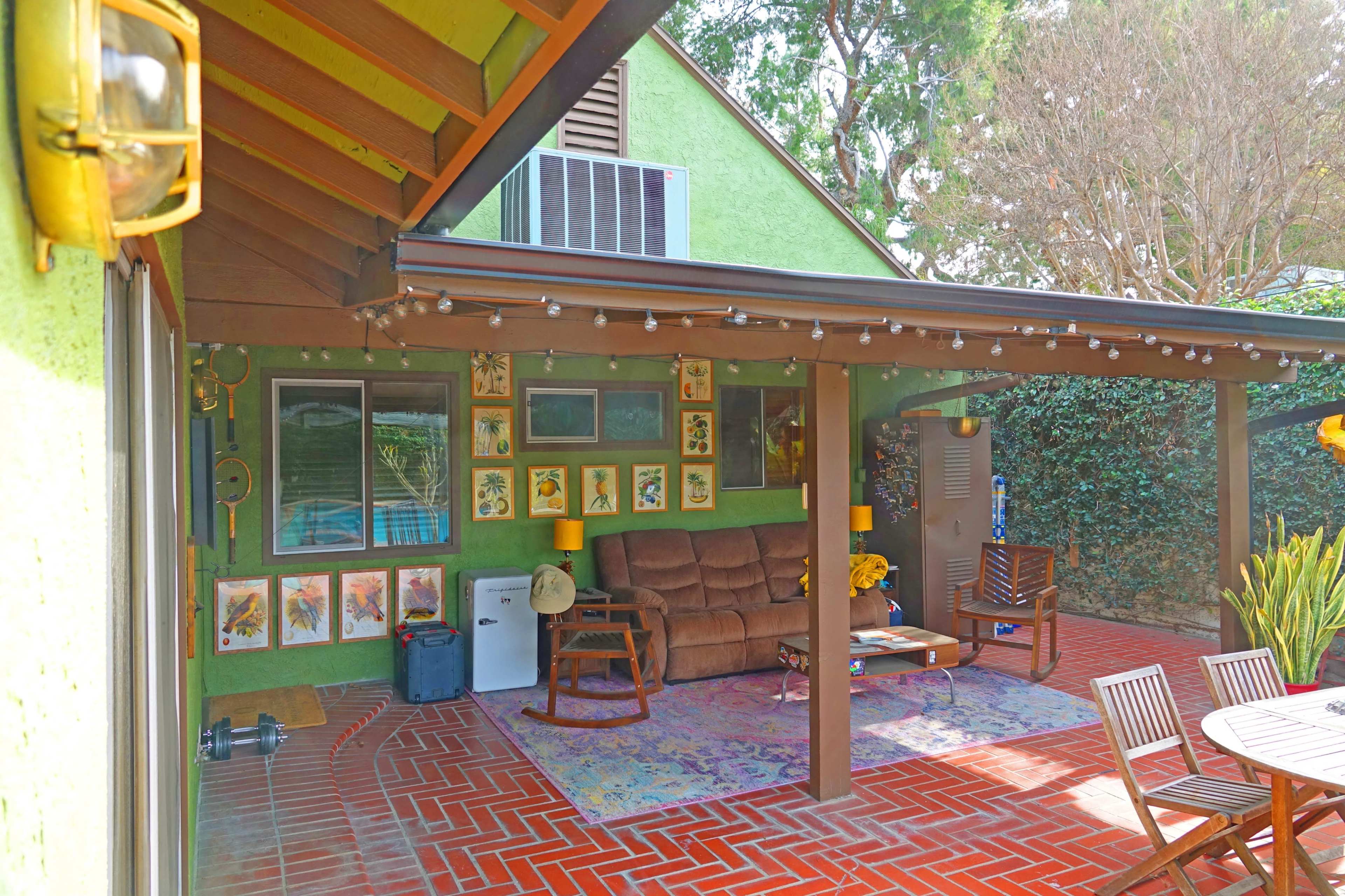The image shows a patio area with a brown couch, a collection of framed art on the walls, and outdoor furniture arranged on a patterned mat.