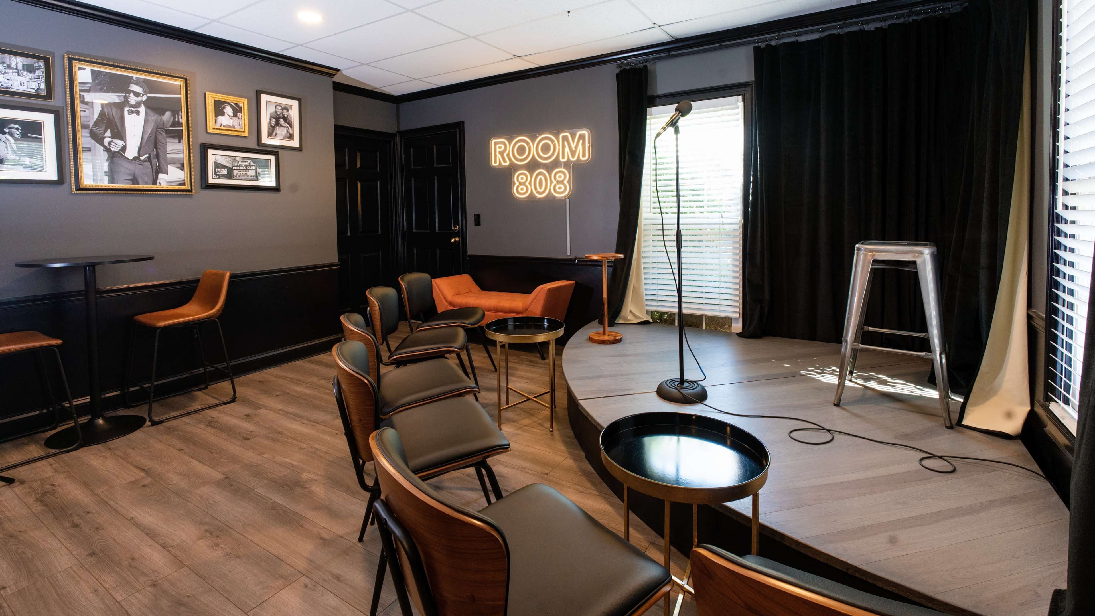The image shows a small performance space with a microphone on a stand, chairs arranged in a semi-circle, and framed photos on the wall, labeled "ROOM 808".