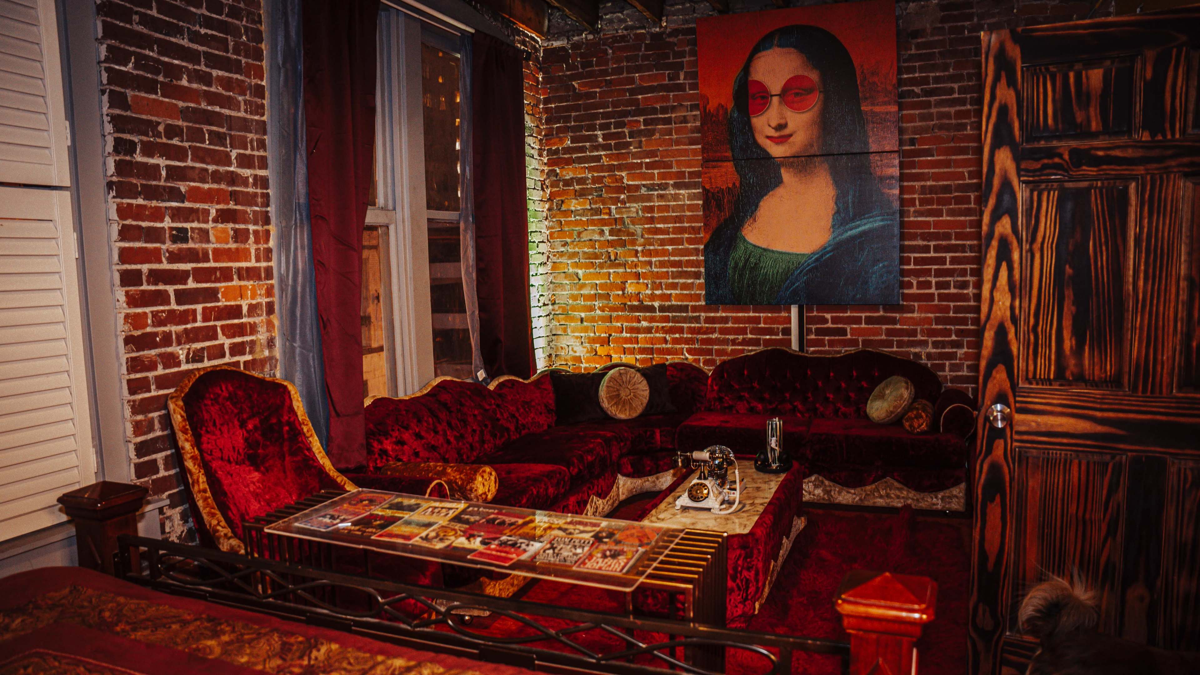 The image shows a cozy living room with red velvet furniture, a glass coffee table displaying magazines, and a large portrait of the Mona Lisa wearing red sunglasses on the wall.