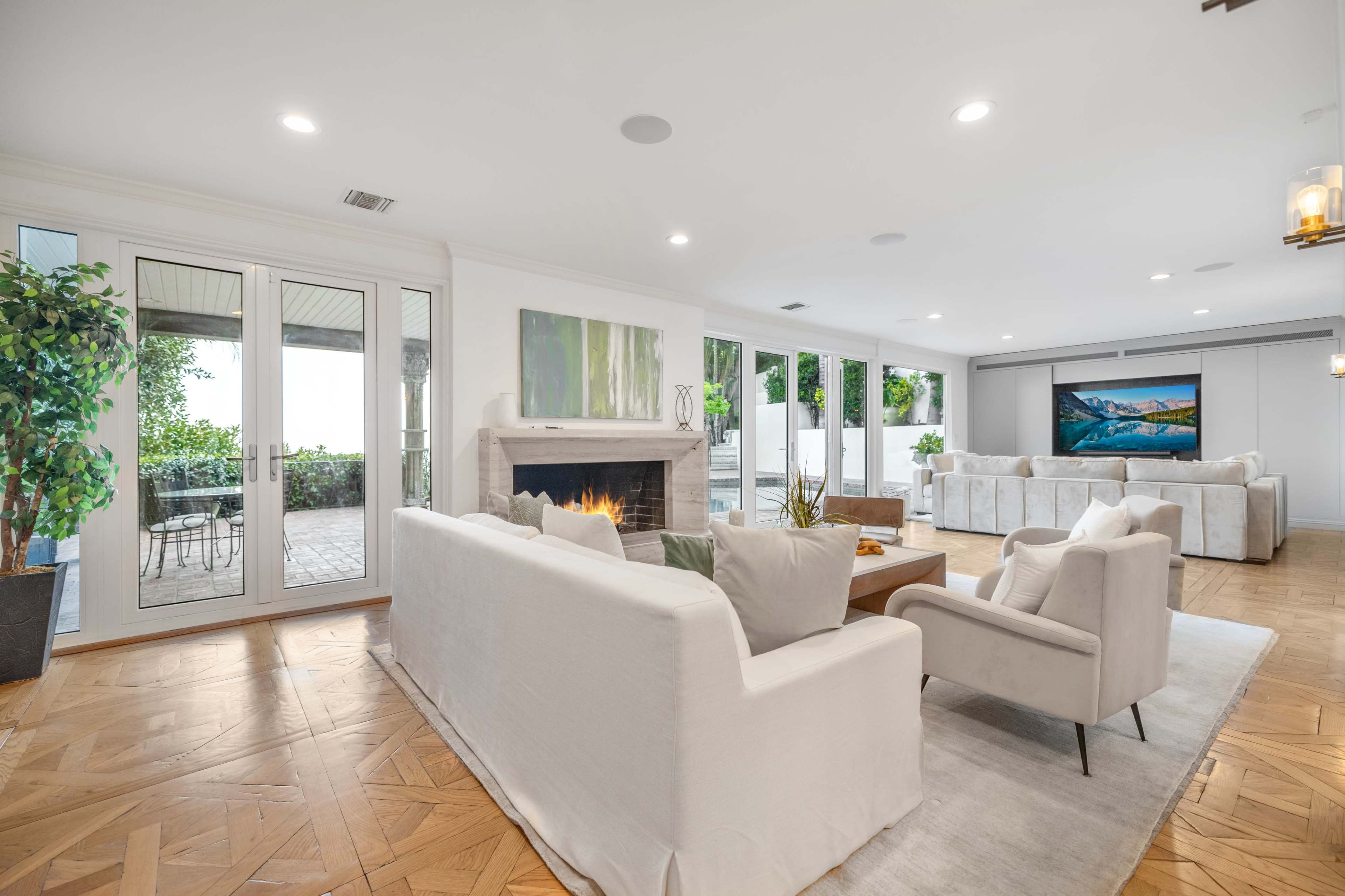 The image shows a spacious living room with a fireplace, large windows, and modern furnishings, featuring a blend of white and neutral tones.