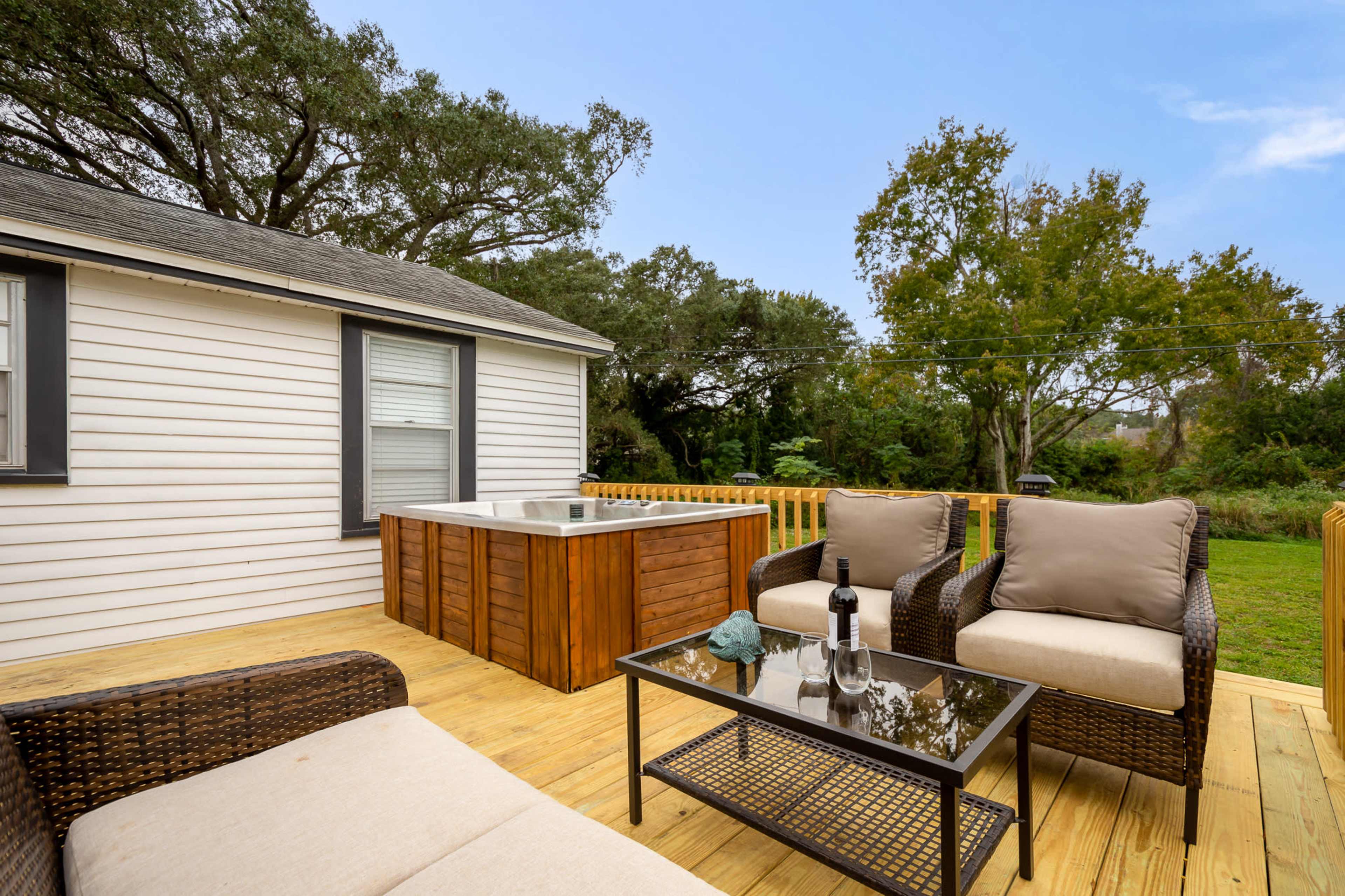 A deck features a seating area with two upholstered chairs and a glass-top table, alongside a hot tub framed by greenery.