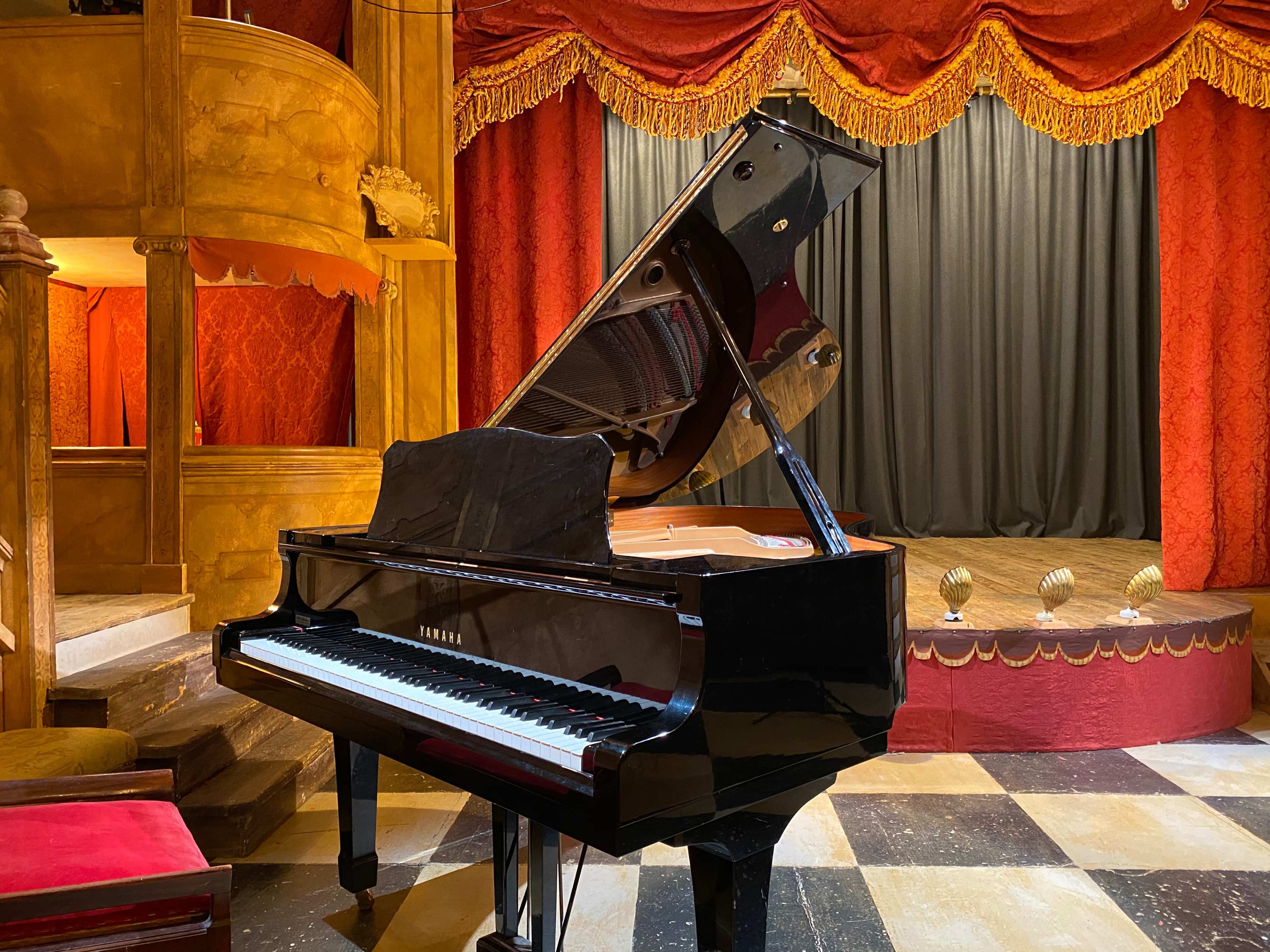A black grand piano sits in front of a velvet-draped stage with gold trim in a theater.