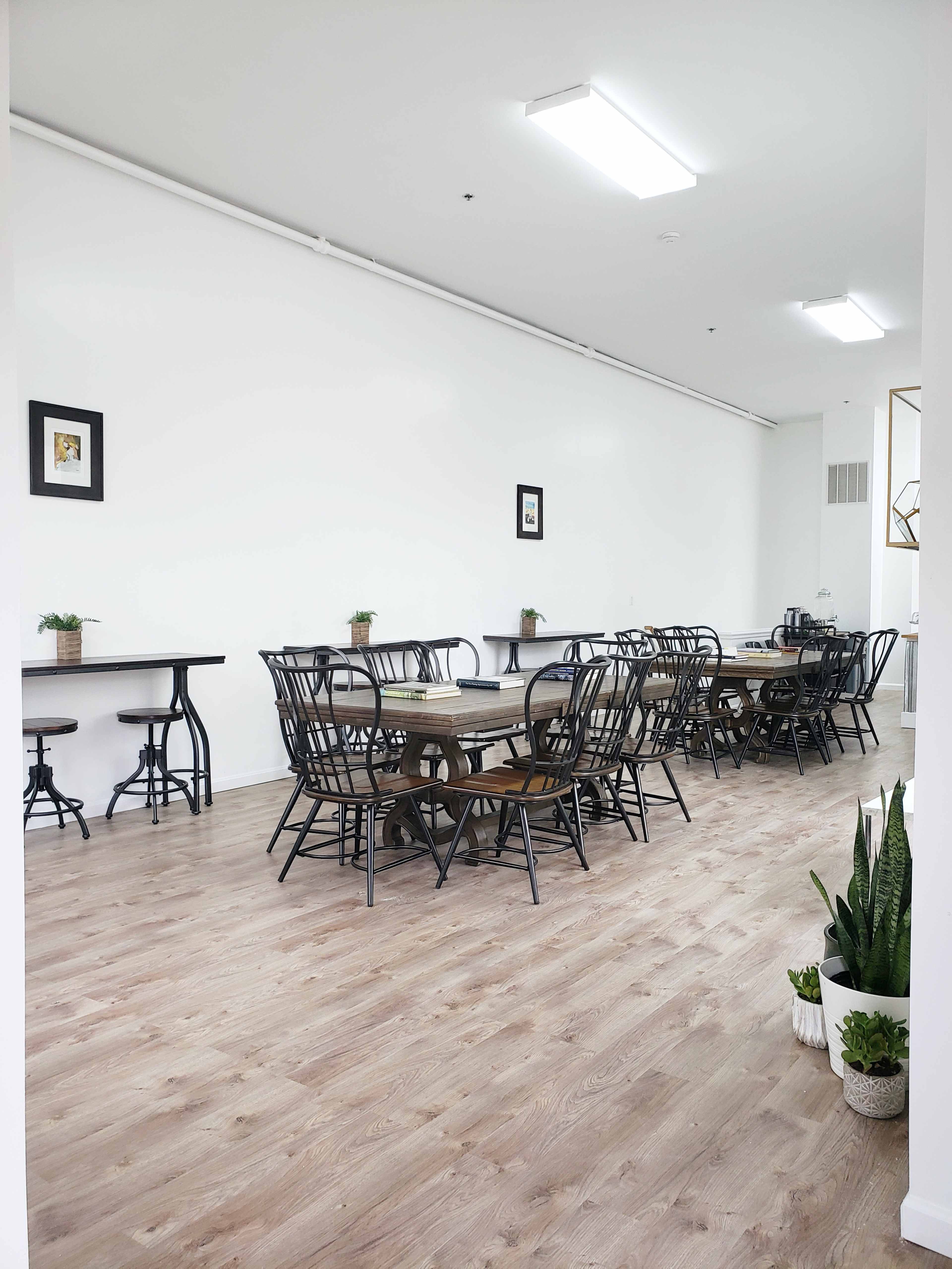 A spacious dining area with multiple tables and black chairs set against white walls and a wooden floor.