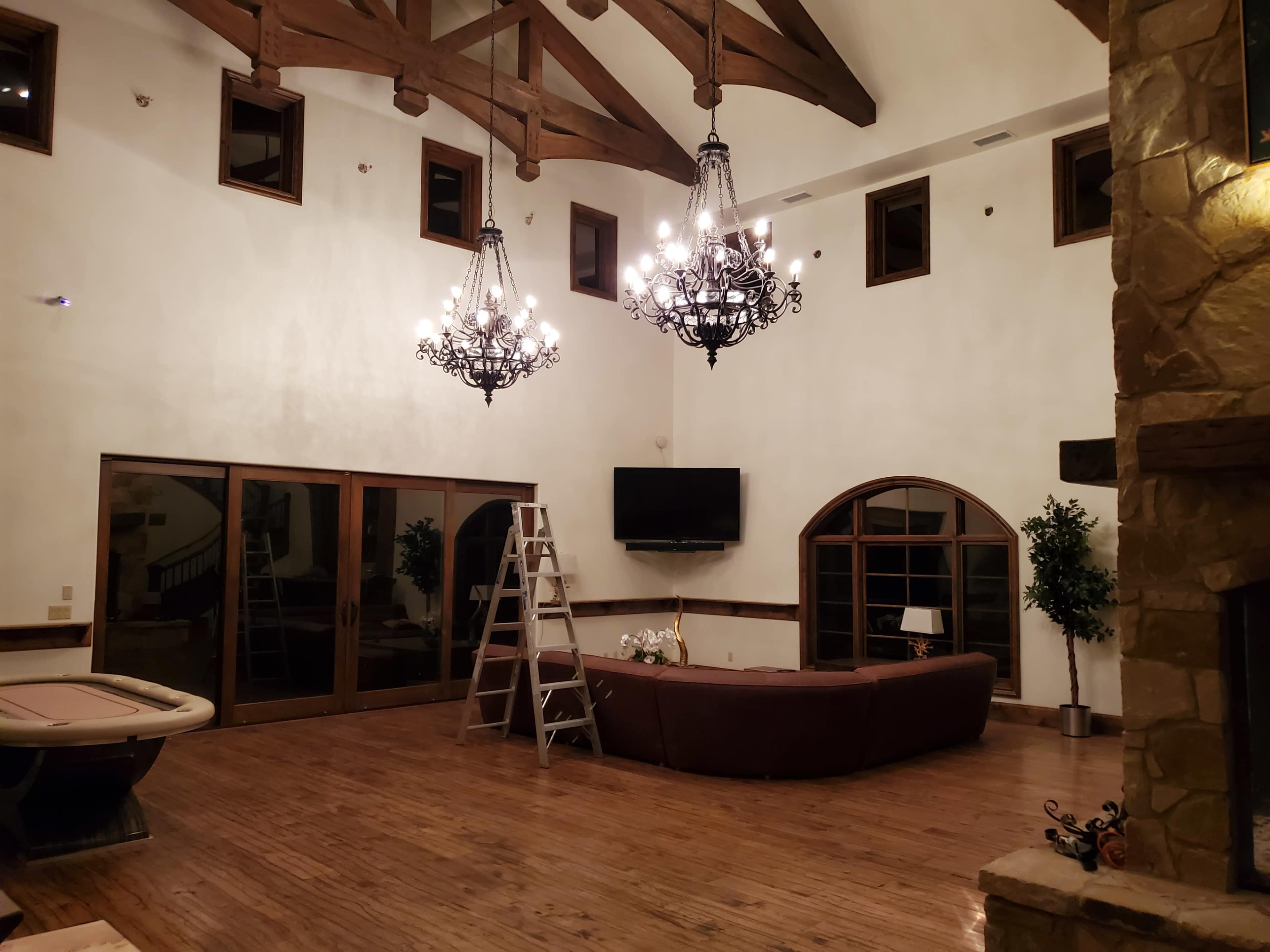 The spacious room features a high ceiling with wooden beams, two ornate chandeliers, and a large sectional sofa with a ladder nearby.
