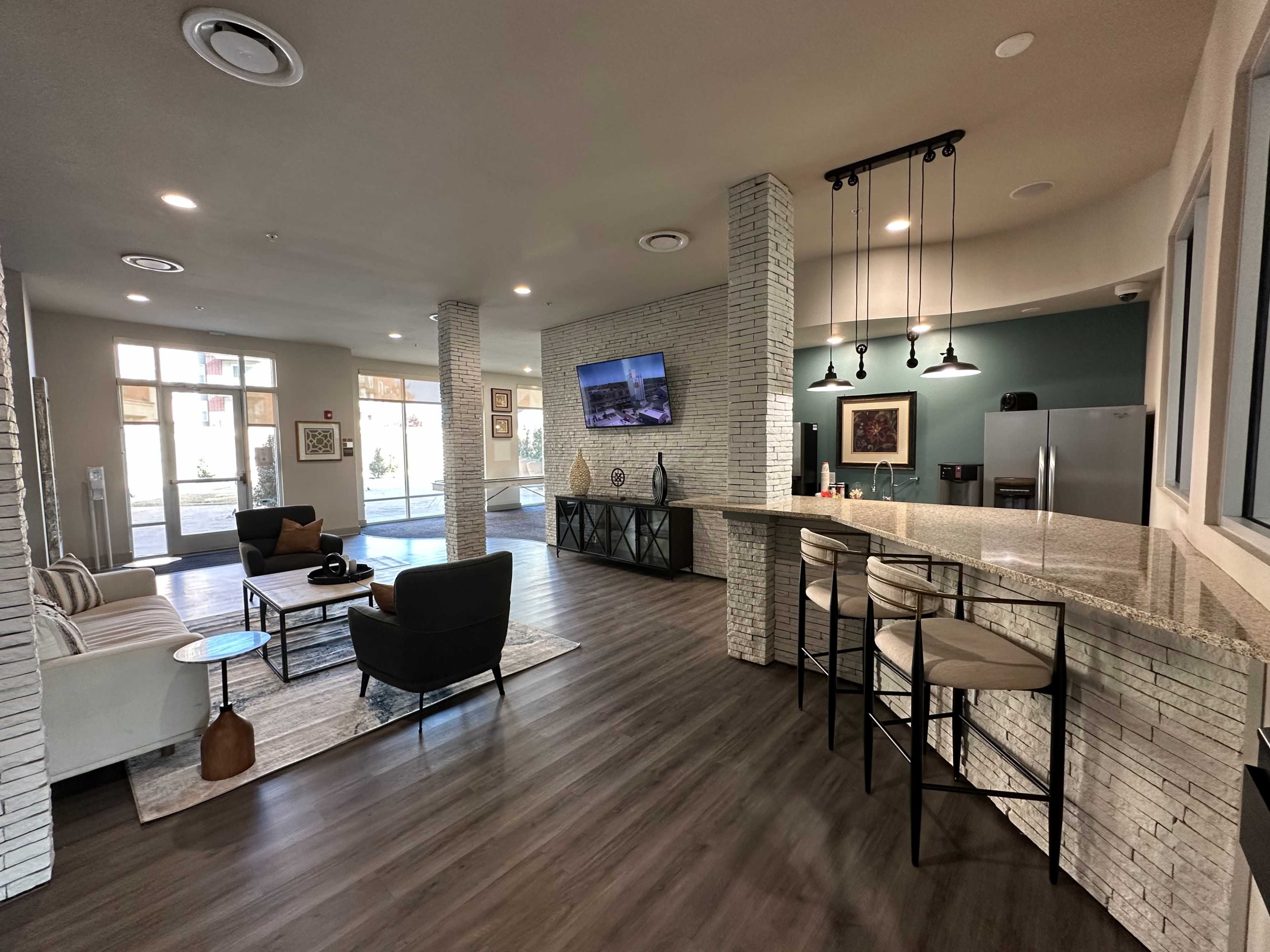 The image shows a modern lounge area featuring a granite countertop bar, seating arrangements, and large windows that allow natural light.