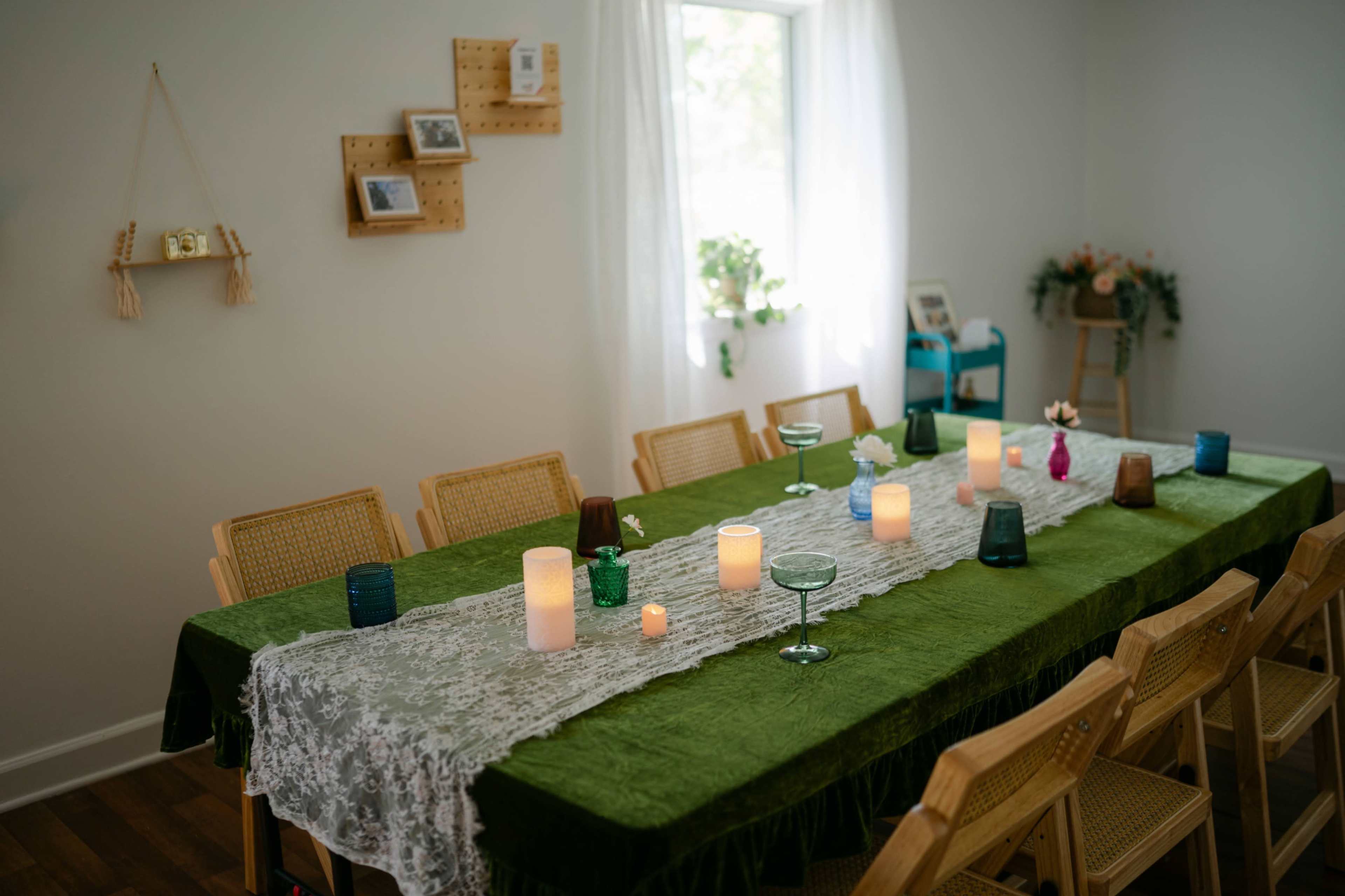 A long dining table is set with green fabric, candles of various sizes, and decorative items, surrounded by wooden chairs in a bright room.